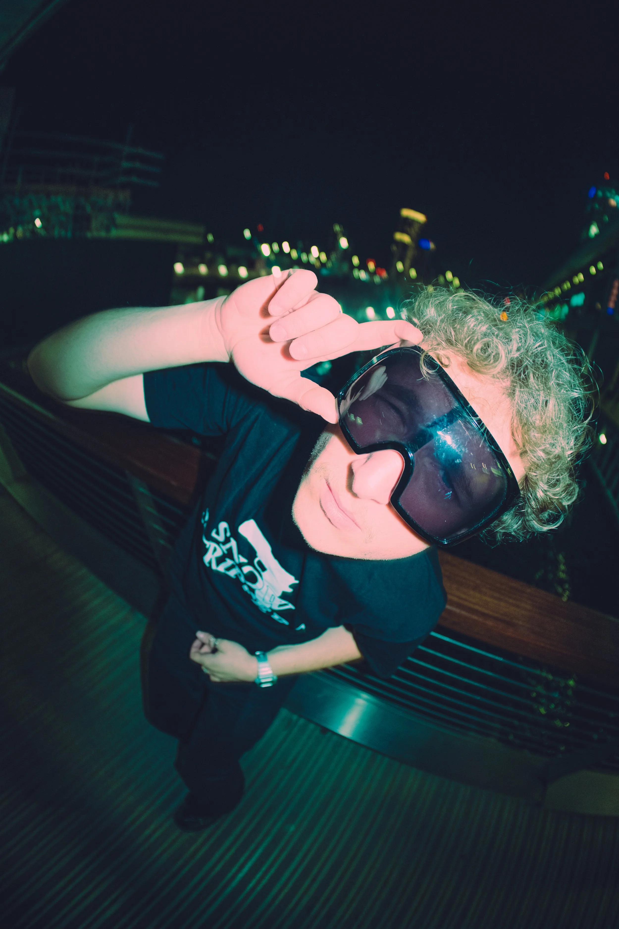 Person with curly blonde hair wearing sunglasses, a black t-shirt, and a watch, making a gesture toward the camera, in an indoor setting with blurred city lights visible through the window.