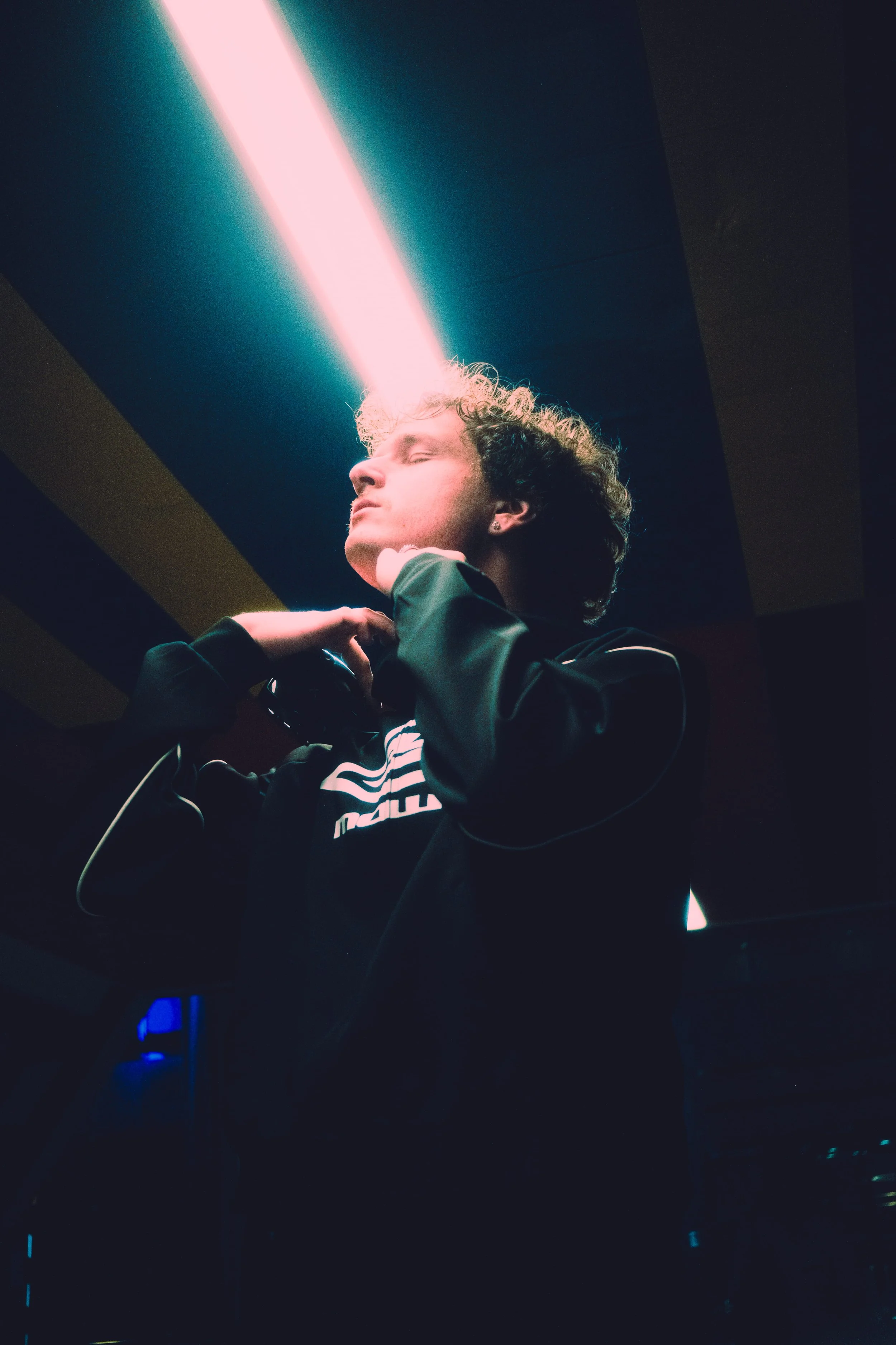 A person with curly light brown hair, wearing a black Nike jacket, is seen with eyes closed, scratching their neck, under a pink neon light in a dark room.