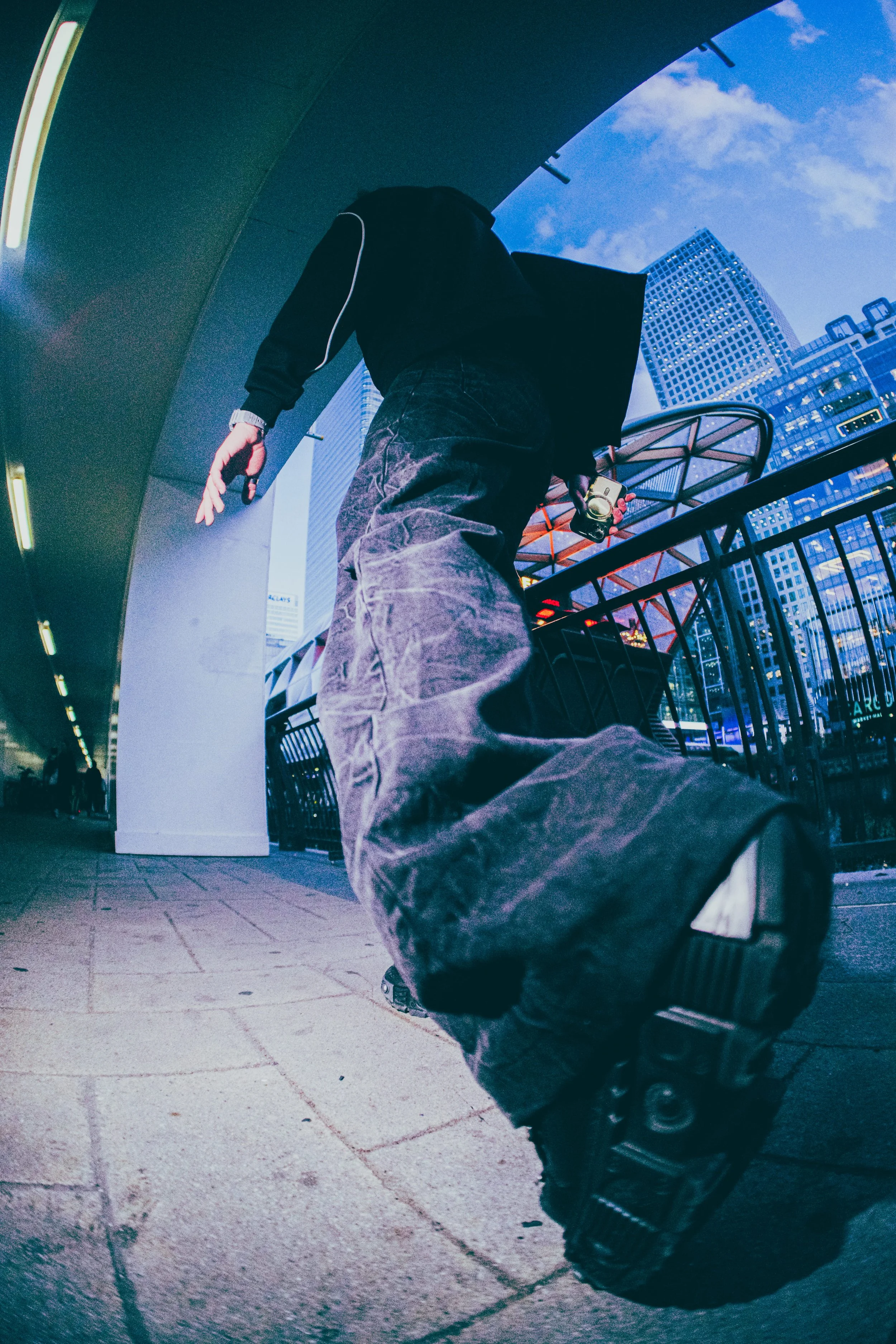 Low-angle view of a person walking on city sidewalk, holding a camera, with tall modern buildings and a partly cloudy sky in the background.