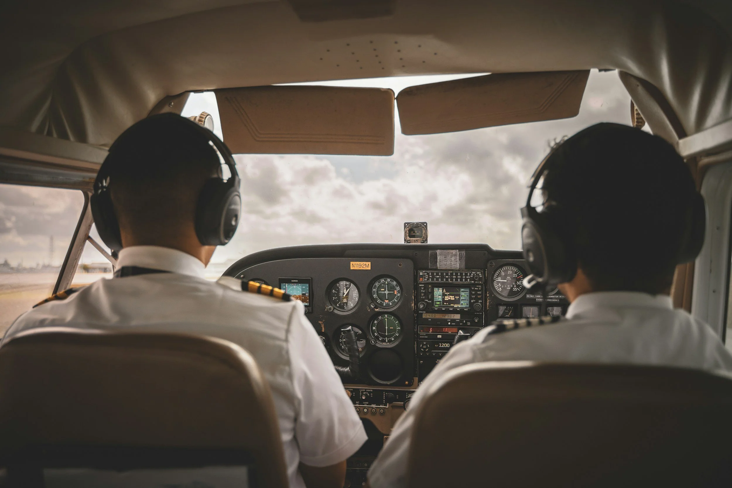 Inside view of an airplane cockpit showing two pilots in uniform wearing headsets, with the instrument panel and controls in front of them and a cloudy sky outside the windshield.