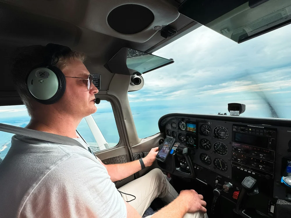 A man flying a small airplane, sitting in the cockpit with headphones and sunglasses, holding a phone with a map app, sky and clouds visible outside the windows.