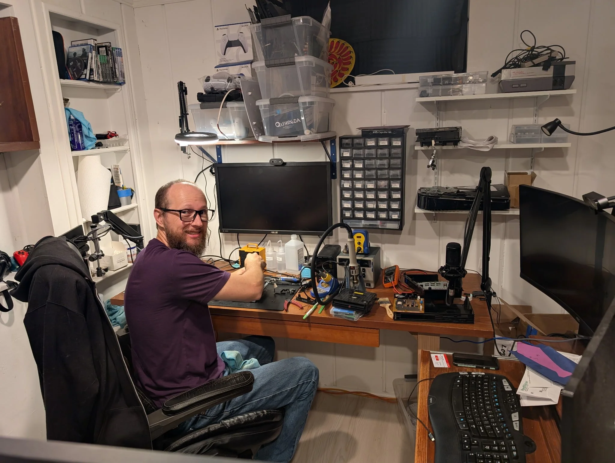 A man with glasses and a beard seated at a cluttered desk in a room, smiling at the camera, surrounded by electronic equipment, tools, and shelves filled with boxes and electronic parts.
