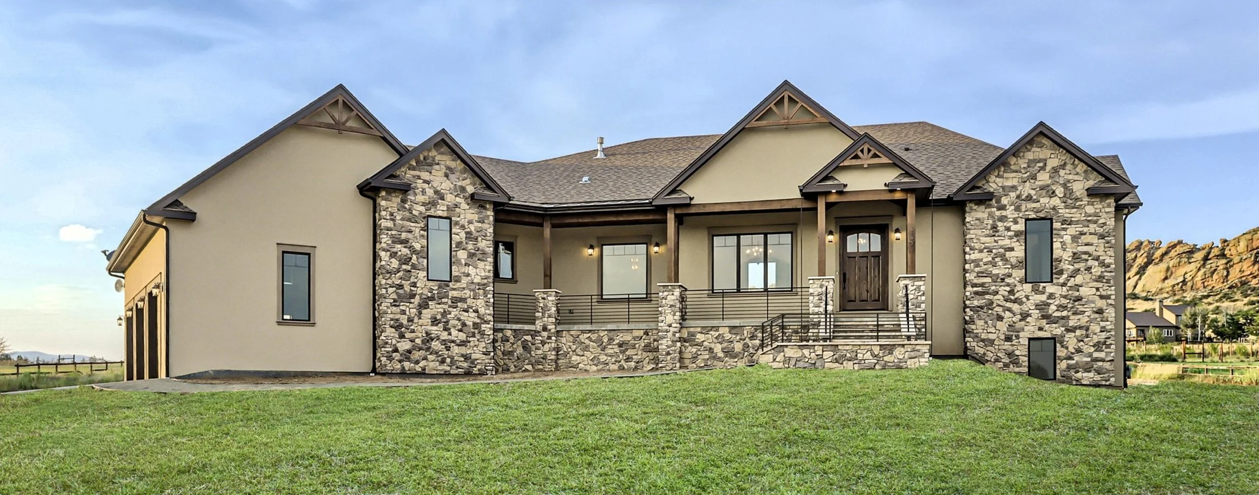 Modern house with stone and stucco exterior, front porch with stairs, large windows, located in a grassy yard with mountains in the background.