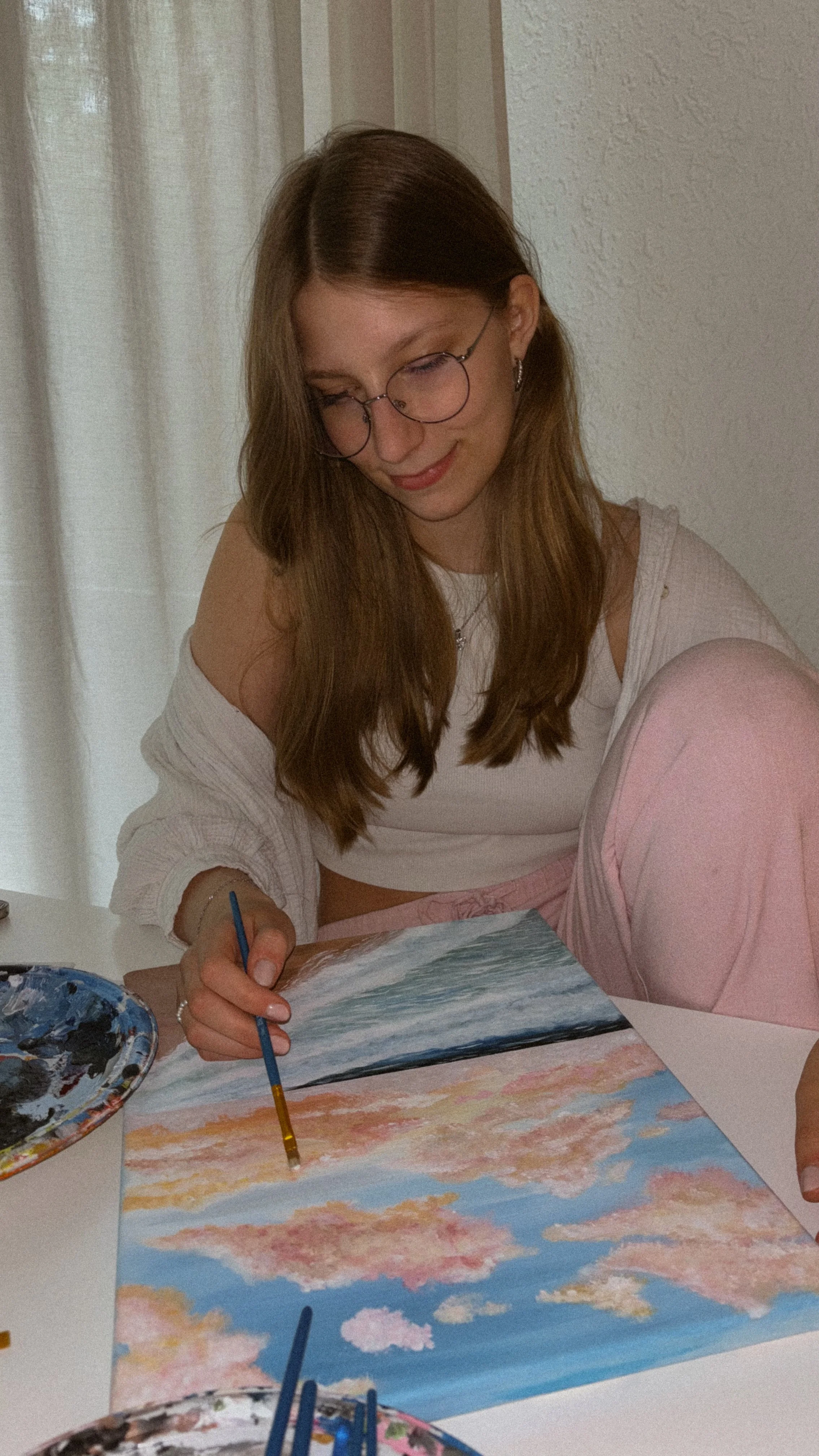 Junge Frau sitzt am Tisch und malt ein Aquarell eines Sonnenuntergangs am Himmel mit Wolken.