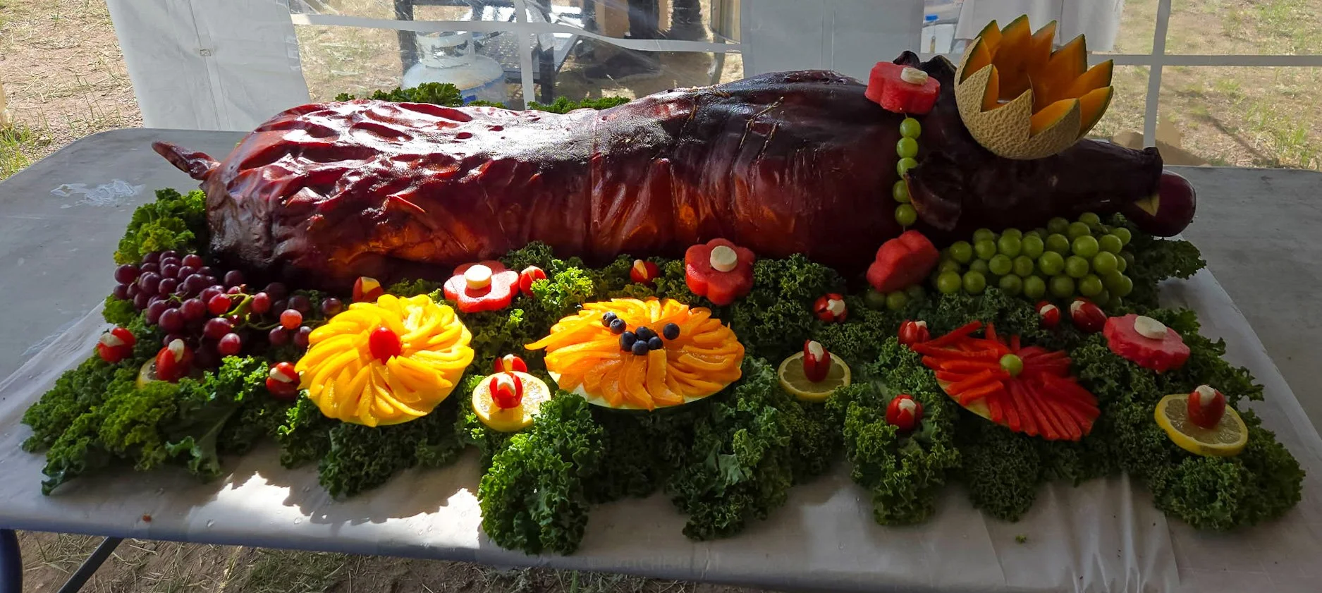 A large, roasted whole pig laid on a bed of leafy greens and surrounded by fruit garnishes. The pig's head is decorated with a carved melon flower, and there are various fruit slices and decorations around the pig, including strawberries, grapes, kiwi, and citrus slices.