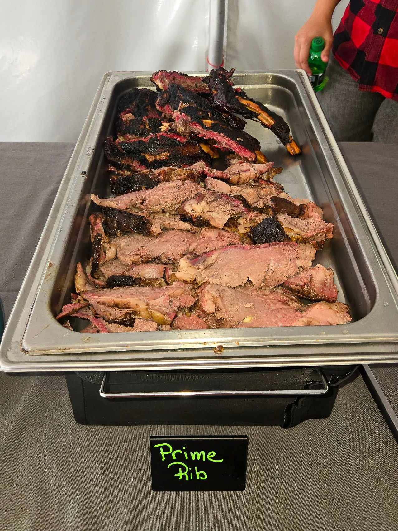 A tray of cooked prime rib with slices of medium-rare and well-done meat, some with a charred crust, on a table with a small black sign labeled "Prime Rib" in green.