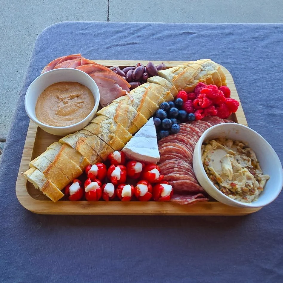 A wooden tray with sliced bread, grapes, berries, sliced meats, cheeses, candy, and bowls of peanut butter and a dip or pâté, arranged on a gray table.