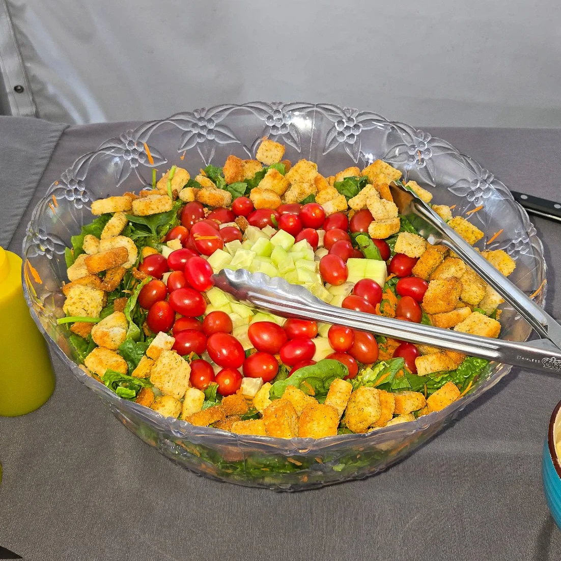 Large glass bowl with a salad containing cherry tomatoes, chopped cucumbers, greens, croutons, and possibly cheese, with a pair of tongs inside.
