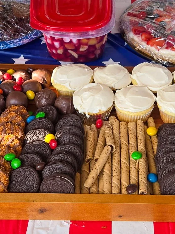 Assorted baked goods and candies on a table, including a variety of cookies, chocolate-covered candies, cupcakes with white frosting, and rolled wafer cookies.