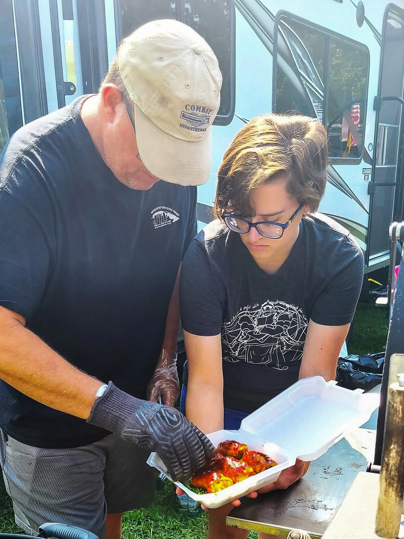 Two people serving food, one holding a tray of baked dishes, outdoors near a black food truck.