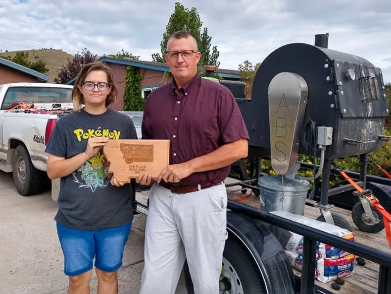 A young girl holding a wooden plaque and a man standing beside her in front of a black smoker grill on a trailer. The girl is wearing glasses and a Pokémon T-shirt. The man is dressed in a maroon shirt and light-colored pants. There is a pickup truck and a rural landscape in the background.