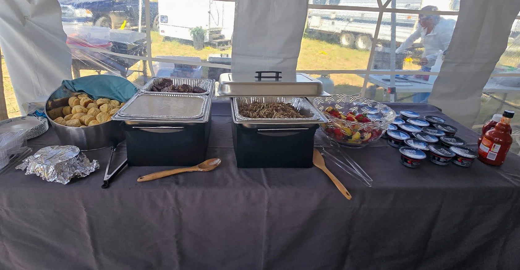 Buffet table with assorted food including meatballs, pasta, fruit salad, yogurt cups, and sauces set inside an event tent.