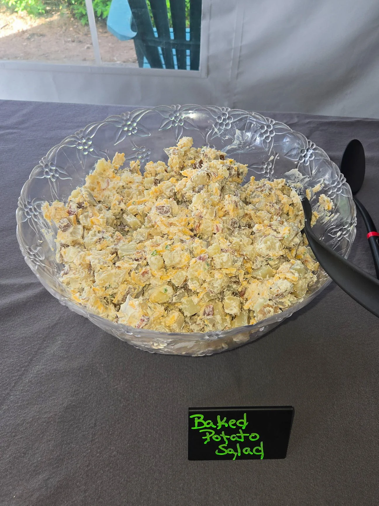A glass bowl filled with baked potato salad on a dark grey table, with a small black sign reading "Baked Potato Salad" in green handwritten text placed in front of the bowl.