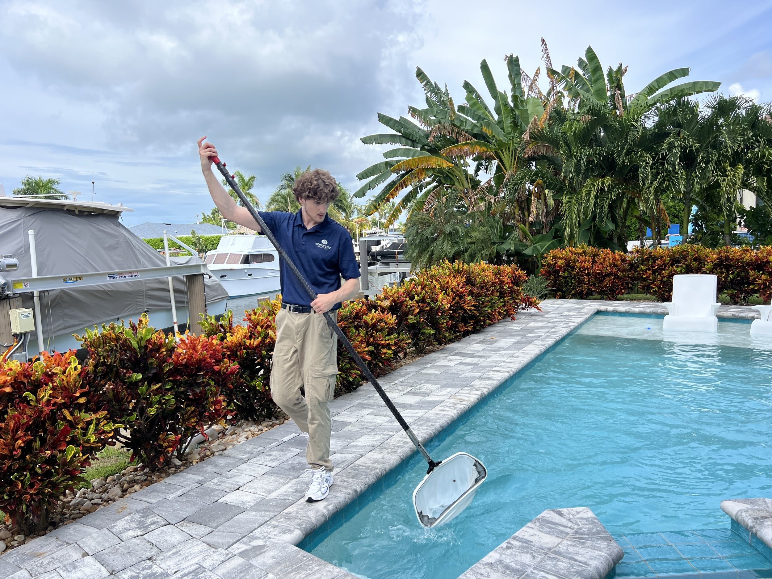 A young man with curly hair wearing a navy blue shirt and khaki pants is standing by a swimming pool holding a pool skimmer net, cleaning debris from the pool water. There are boats and lush green plants in the background, along with a partly cloudy sky.