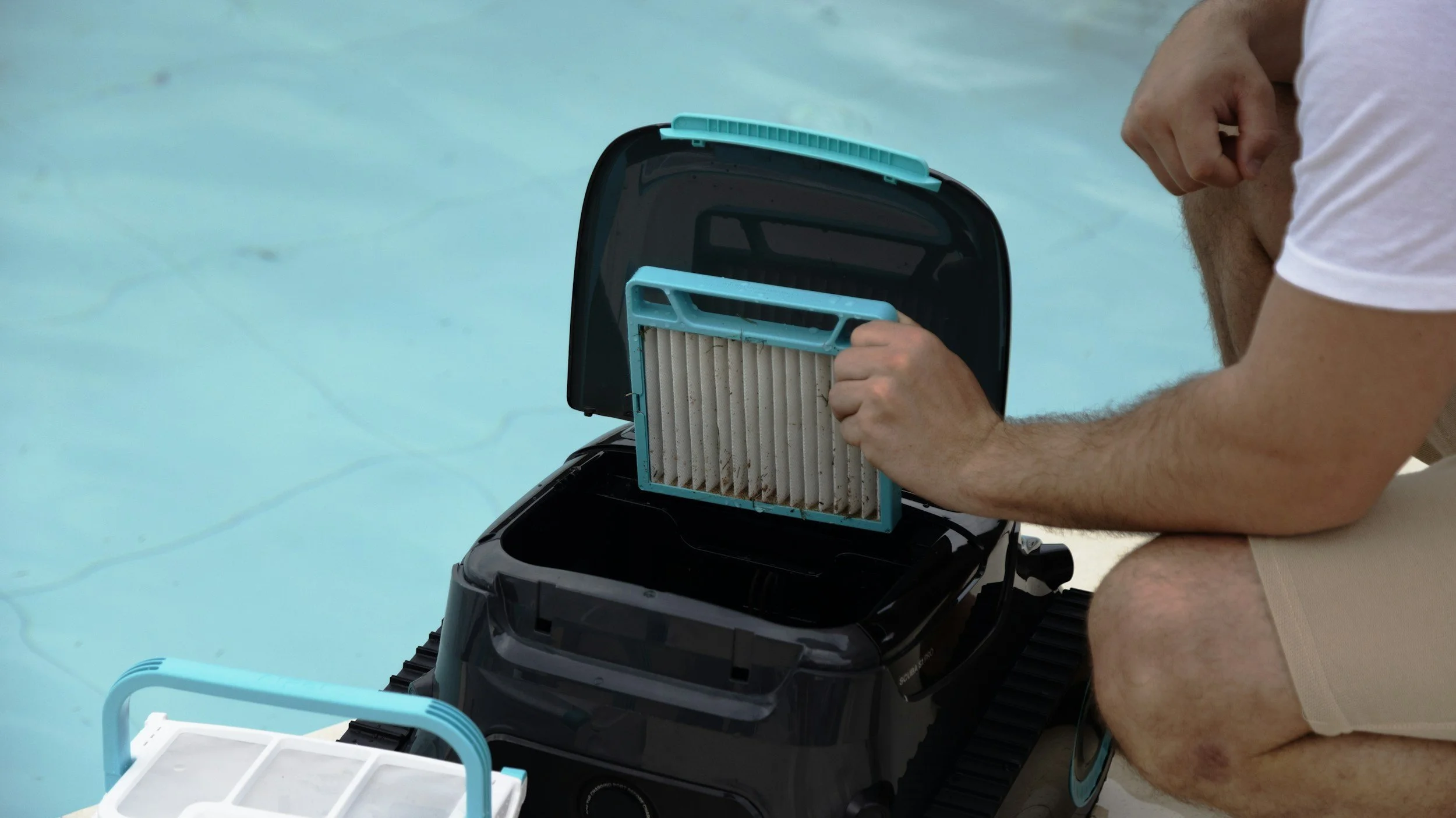 A person is kneeling beside an outboard motor, removing a dirty air filter from the motor, near a swimming pool.