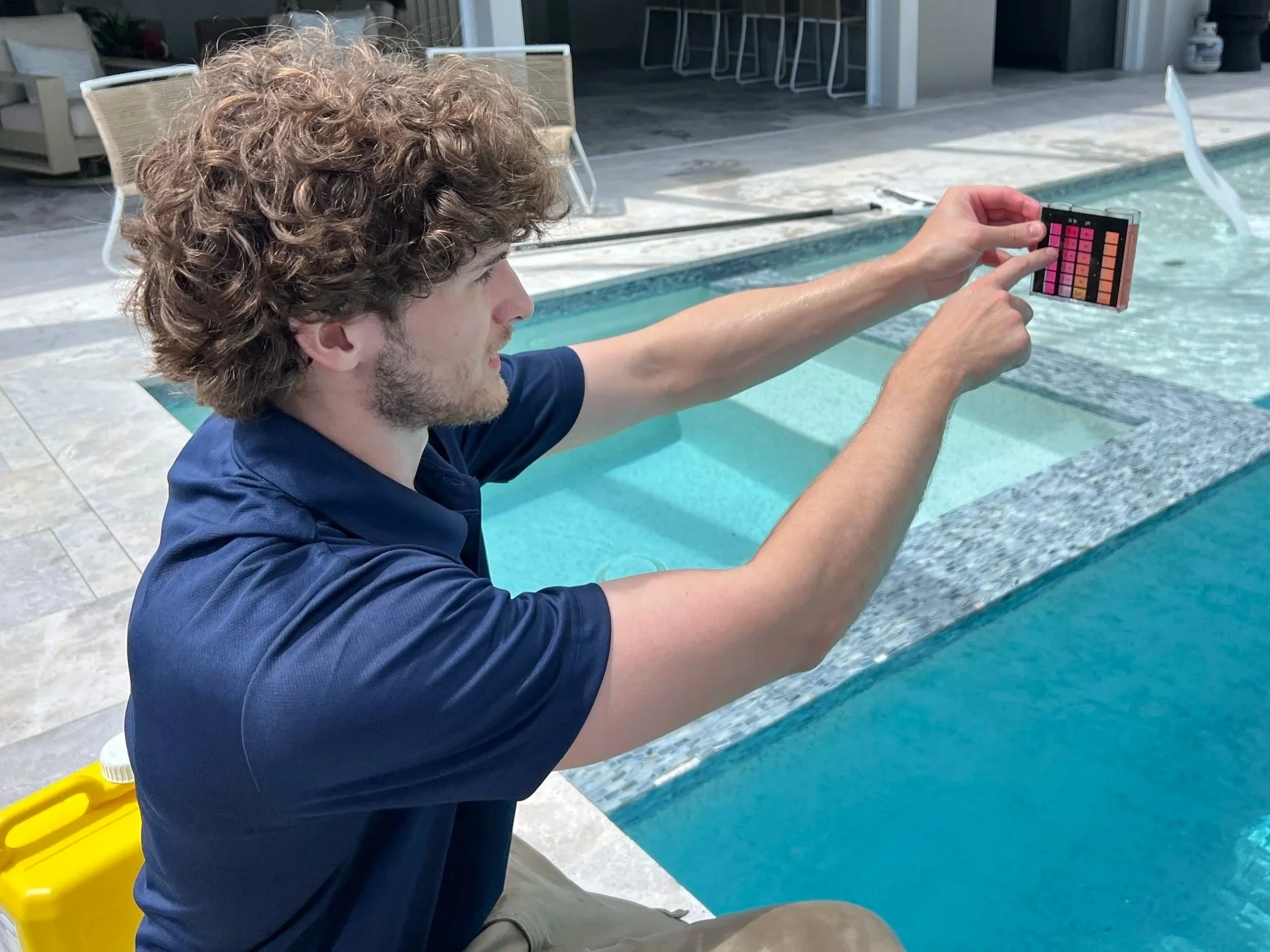 A young man with curly hair and a beard, wearing a dark blue shirt, is sitting by a swimming pool. He is holding a color palette or testing kit with pink and orange shades, extending his arm over the pool.