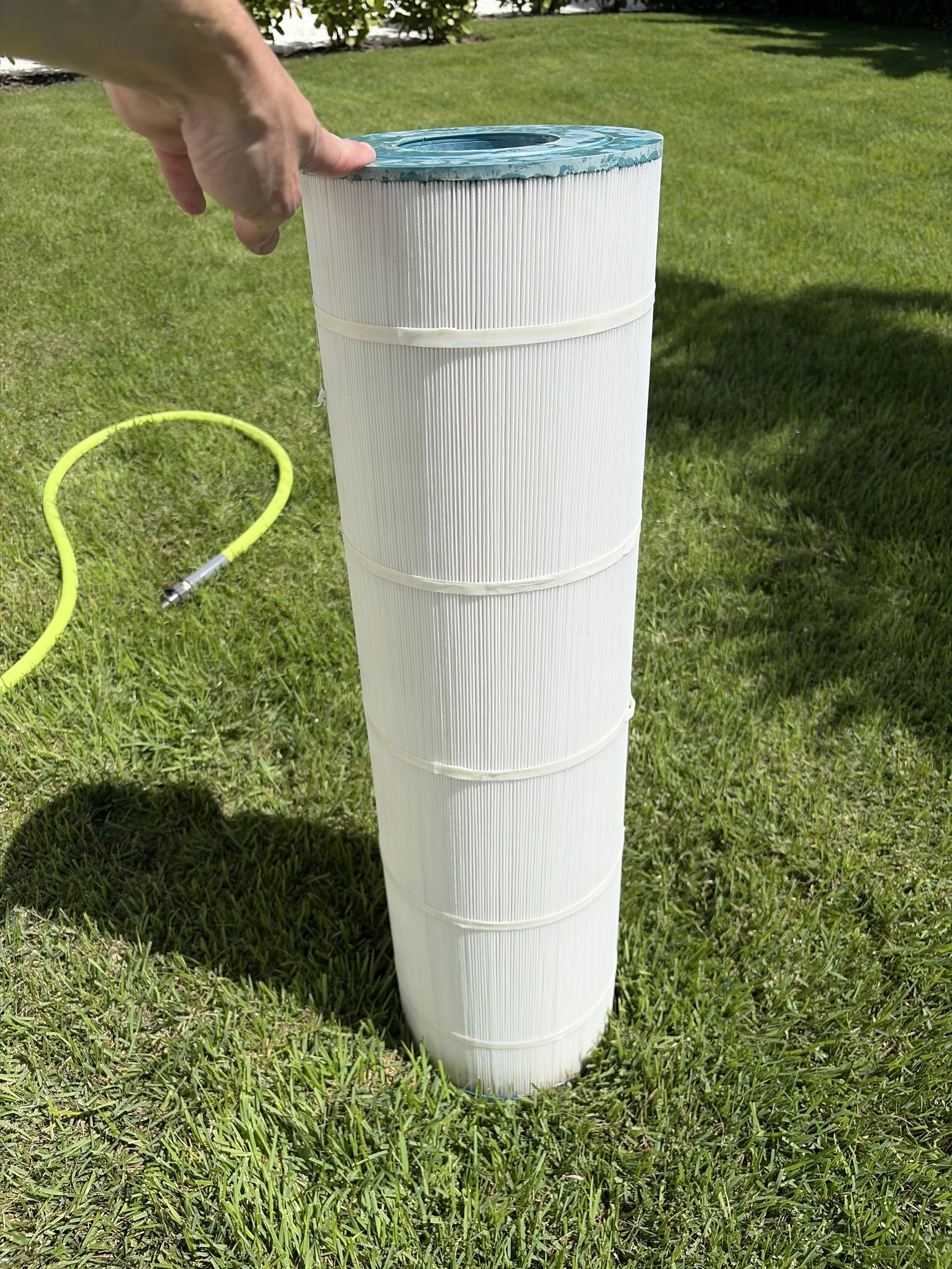 A hand holding a large, cylindrical white pool filter with a ribbed surface, positioned on green grass outdoors with a garden and trees in the background, and a yellow hose nearby.