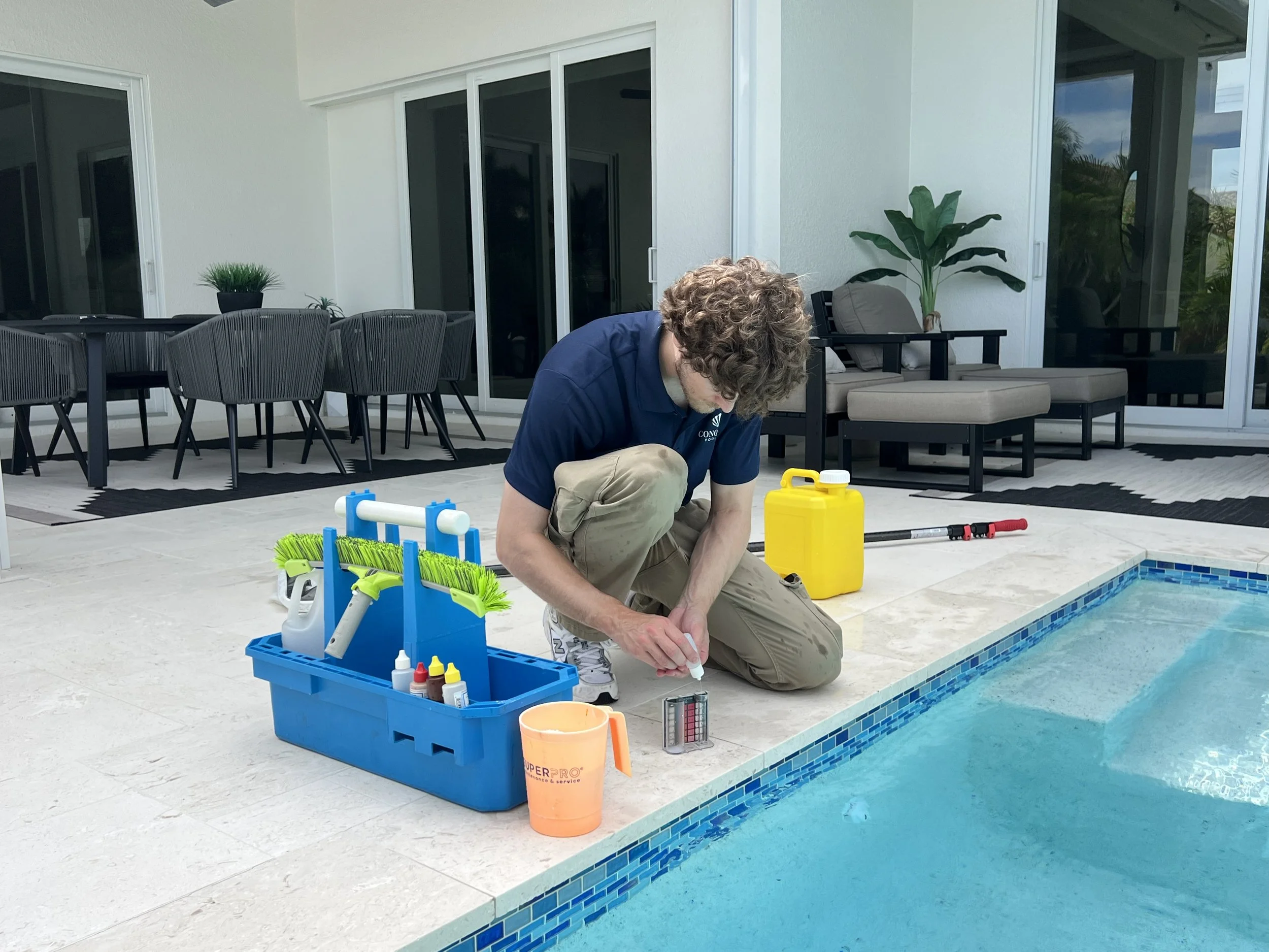 A man kneeling by a swimming pool, preparing to test the water. He is surrounded by pool supplies, including a blue toolbox with various bottles, a yellow container, and testing strips. The background features patio furniture, potted plants, and glass doors leading to a house.