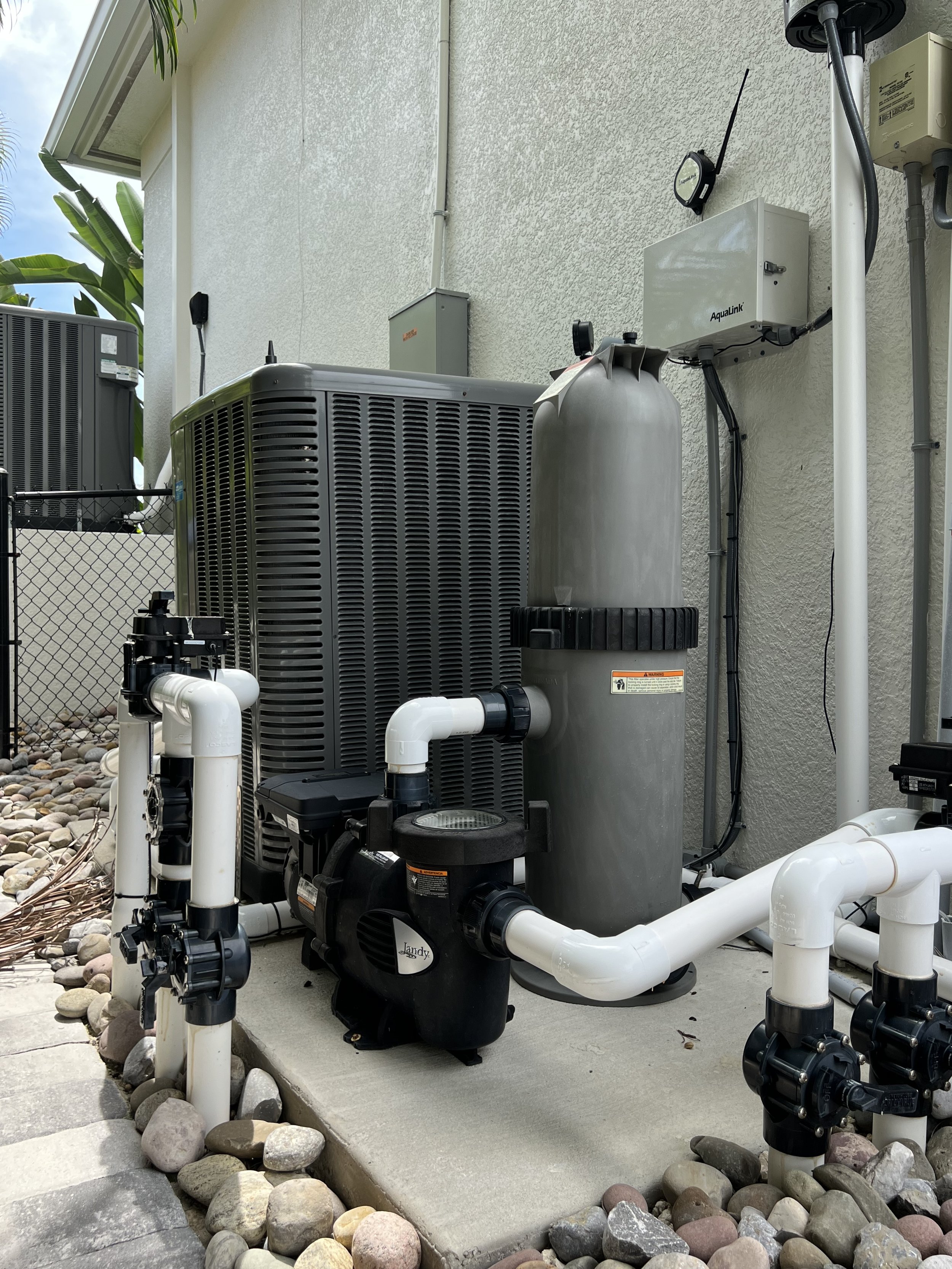Outdoor view of a pool equipment area with pumps, filters, and pipes against a textured wall, with rocks and concrete surface in the foreground.