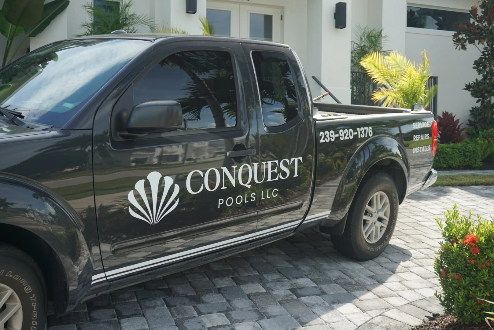Black pickup truck with white and gray branding for Conquest Pools LLC, parked on a paved driveway in front of a modern house with landscaping.