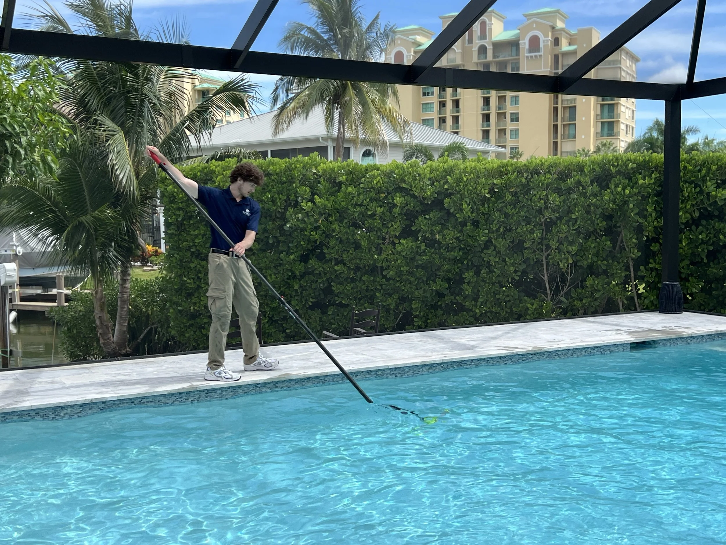 A young man with curly brown hair, wearing a navy blue polo shirt and beige pants, is cleaning a swimming pool using a pool skimmer pole. The pool has clear blue water and a tiled edge. In the background, there are tall palm trees, lush green bushes, and a white house with a metal roof. Modern beige high-rise buildings are visible further back. The scene takes place outdoors on a sunny day with a blue sky.