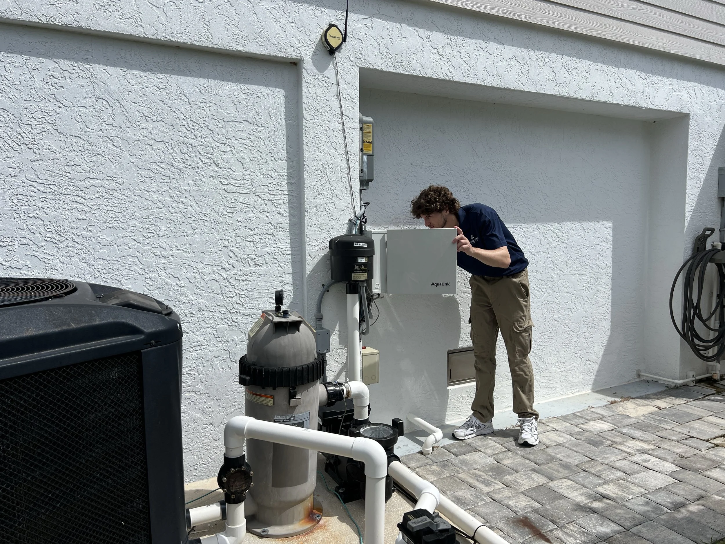 A man with curly hair wearing a navy blue polo shirt and beige pants is working on a water treatment or filtration system outside a white stucco building. He is hunched over near a control panel, with pipes and equipment nearby, including a pool chlorinator and a large black air blower.