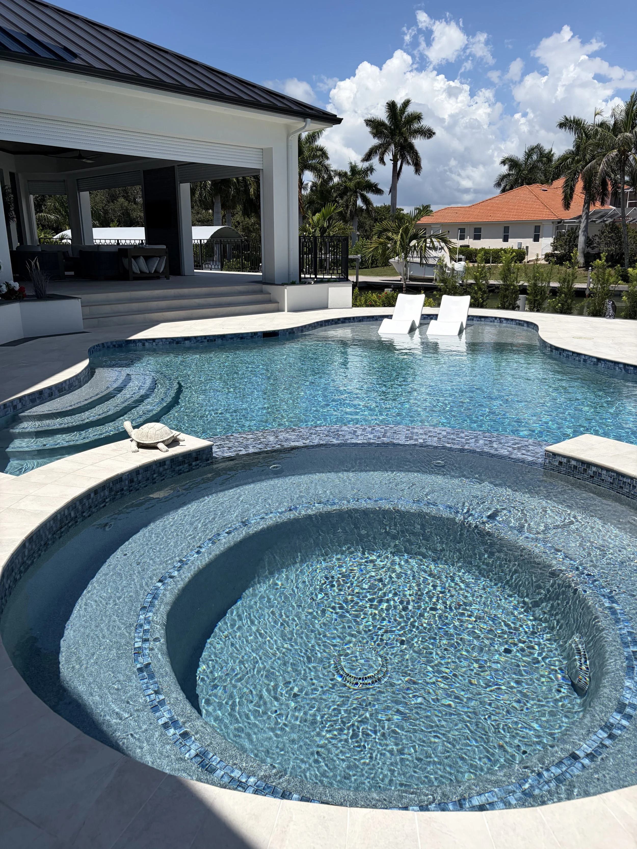 A backyard swimming pool with a small underwater seating area and two white lounge chairs, surrounded by a tiled deck and tropical palm trees under a partly cloudy blue sky.