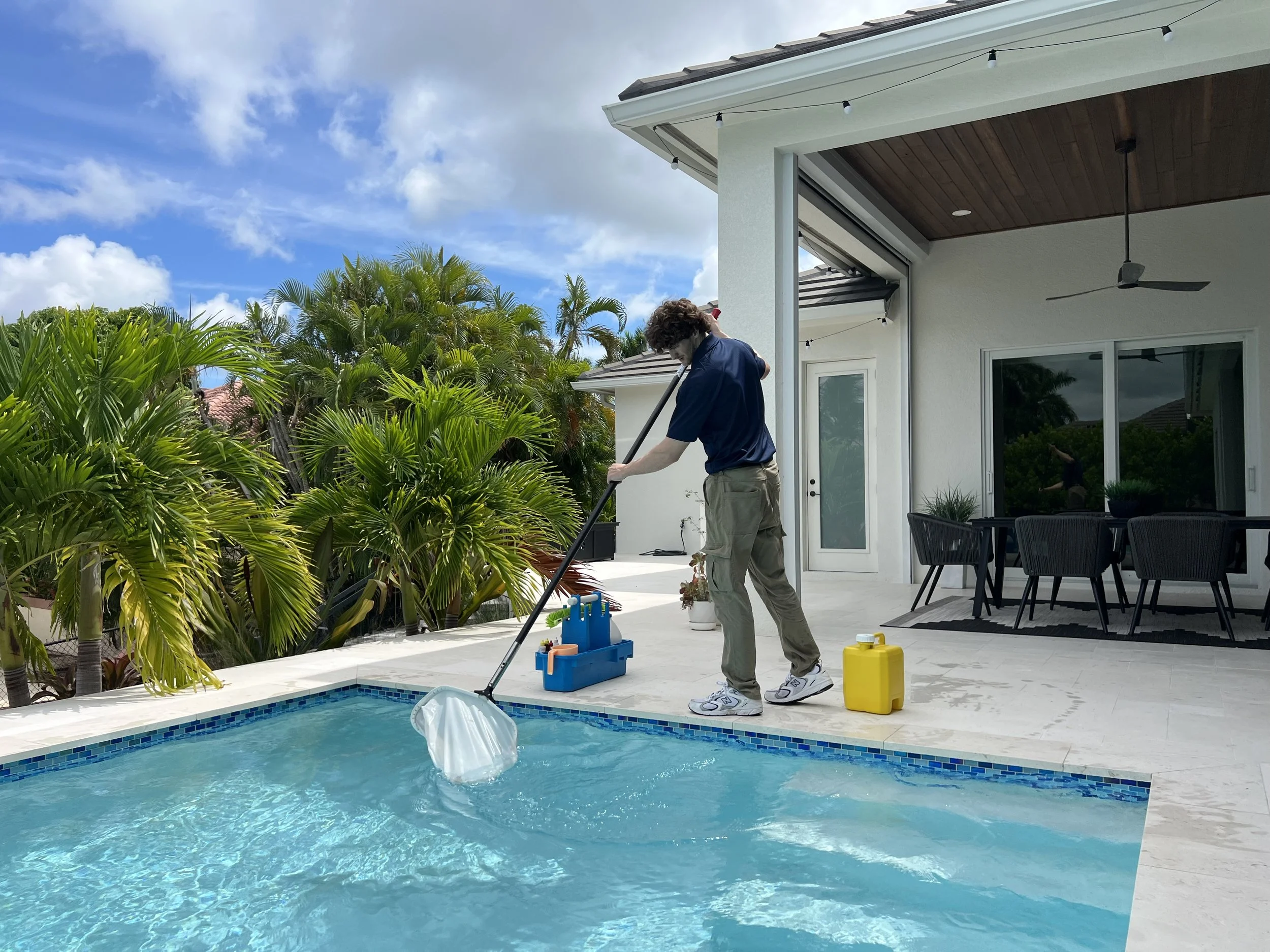 Man in blue shirt and khakis cleaning a backyard swimming pool with a net, surrounded by green plants, a patio with outdoor furniture, and a white house with a covered porch and string lights on a partly cloudy day.