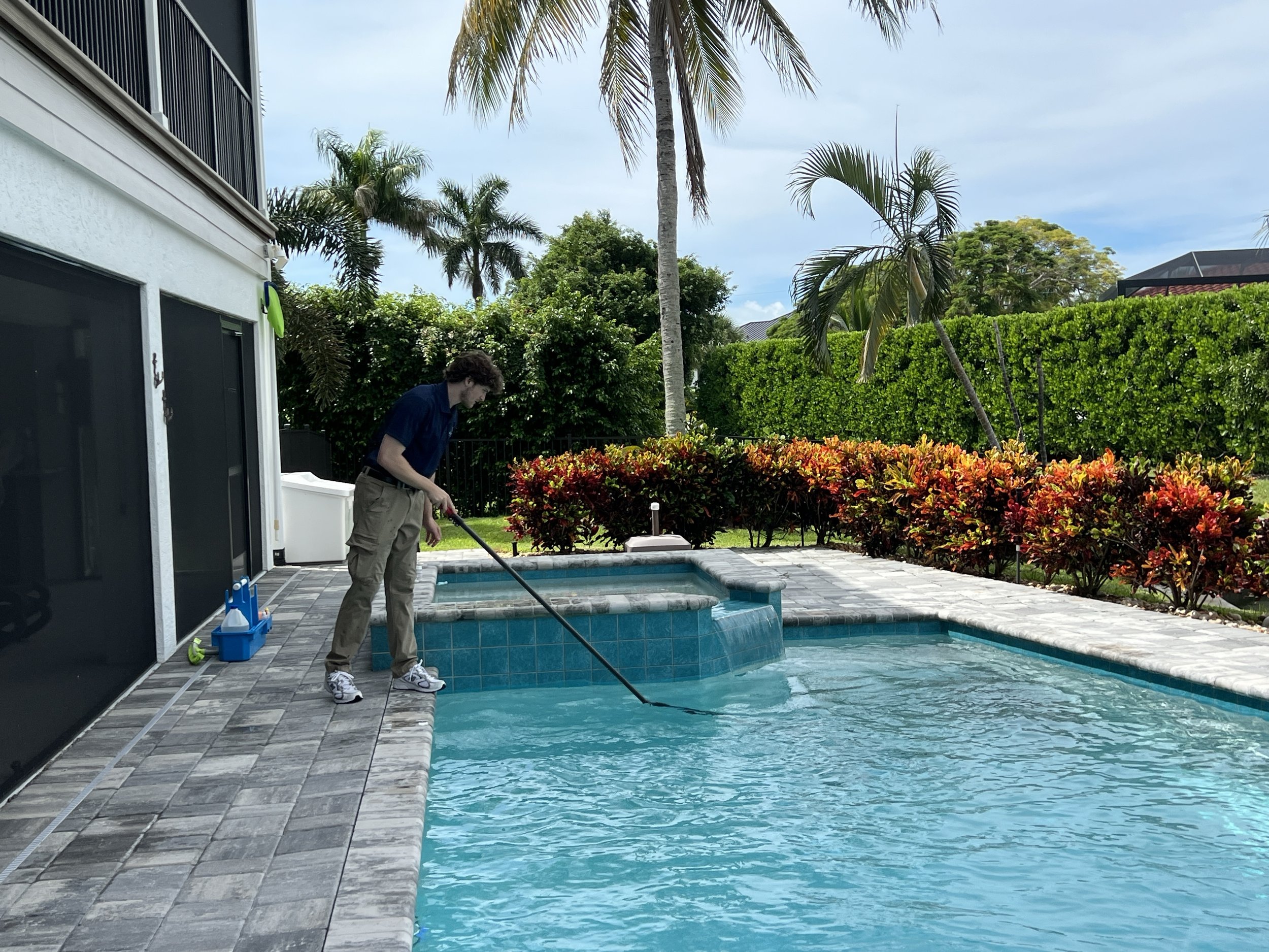 A man cleaning a backyard swimming pool with a net skimmer, next to a house with a white wall, surrounded by tropical plants and trees, under a partly cloudy sky.