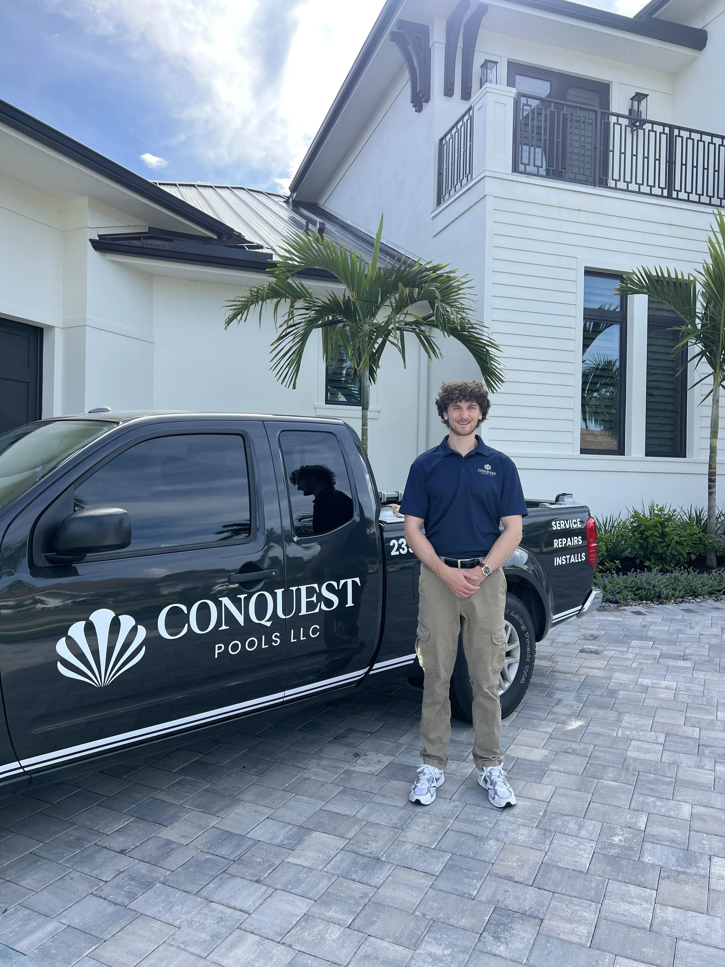 A man in a navy polo shirt and beige cargo pants standing next to a black Conquest Pools LLC service truck in front of a modern white house with black accents and palm trees.
