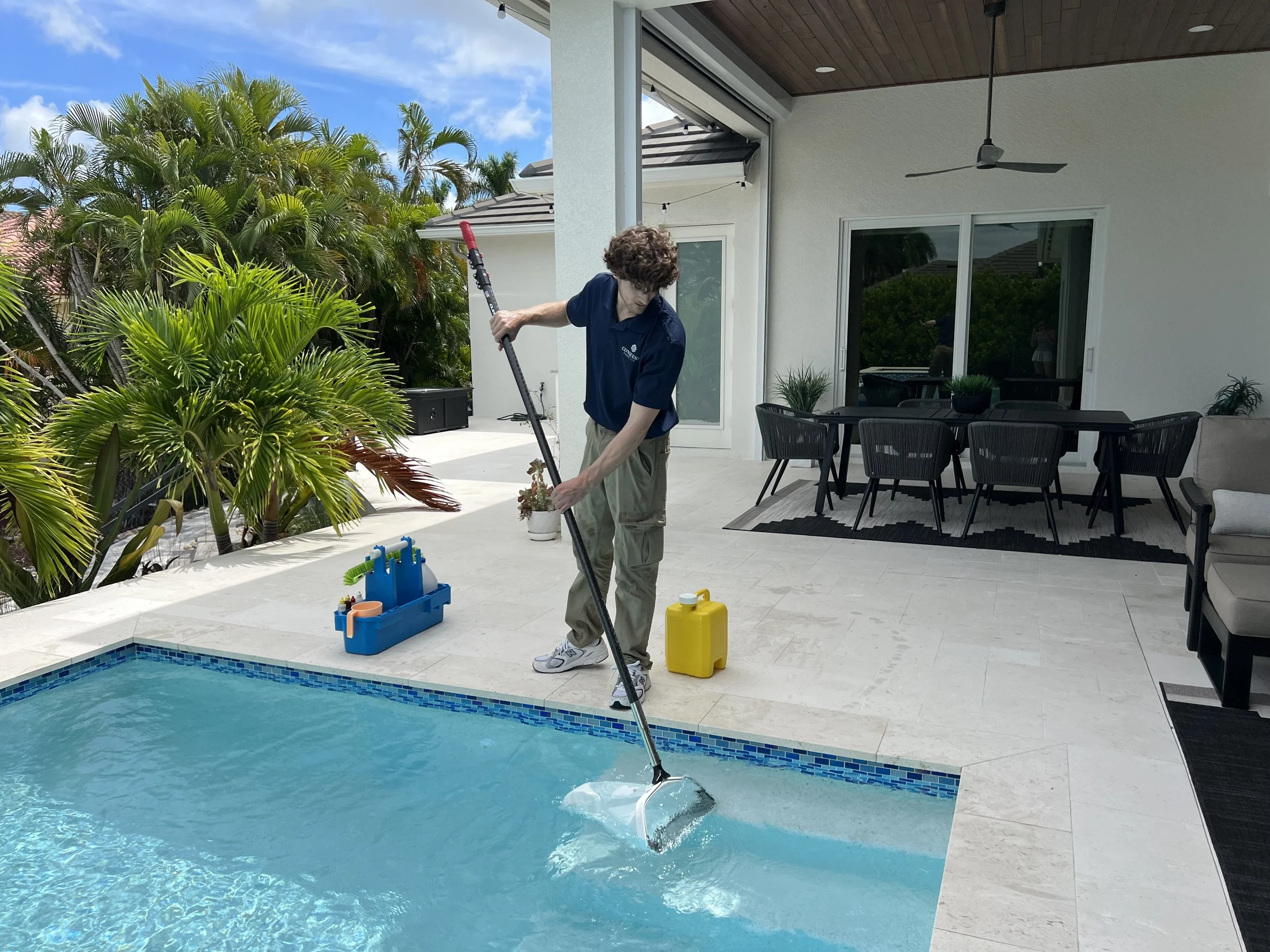 A person cleaning a swimming pool with a long pole, near a patio with outdoor furniture and plants, under a blue sky.