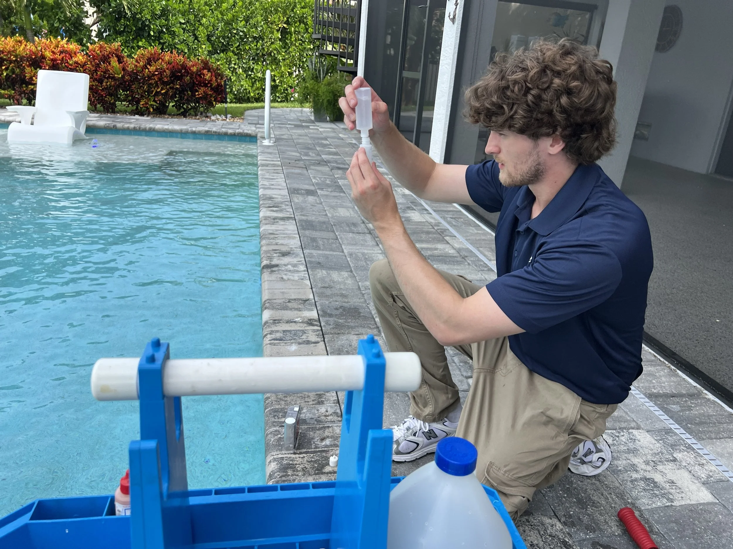 A young man with curly hair and a beard kneeling beside a swimming pool, holding a sterile dropper or pipette, with a pool chemical testing kit, and colorful bushes in the background.