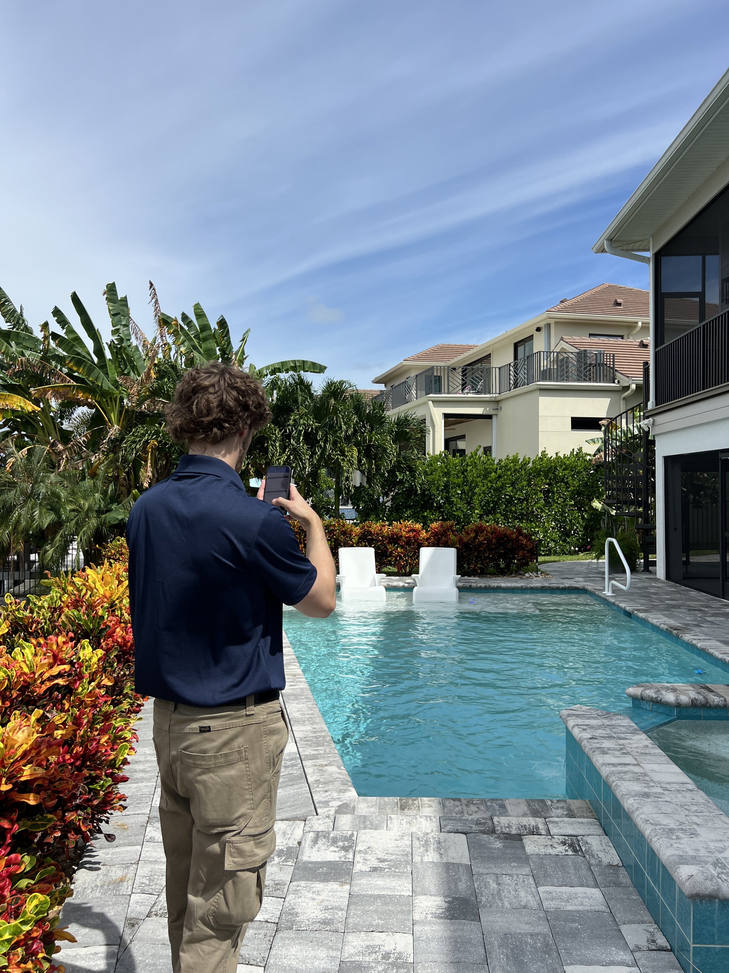 A man with brown curly hair, wearing a navy blue shirt and beige cargo pants, stands by a backyard swimming pool during daytime, taking a photo with his smartphone. The pool is surrounded by a stone tile deck, colorful landscaping, and has slide fixtures at the far end. In the background, there are two-story white residential buildings with balconies, and lush green trees and plants, under a clear blue sky.