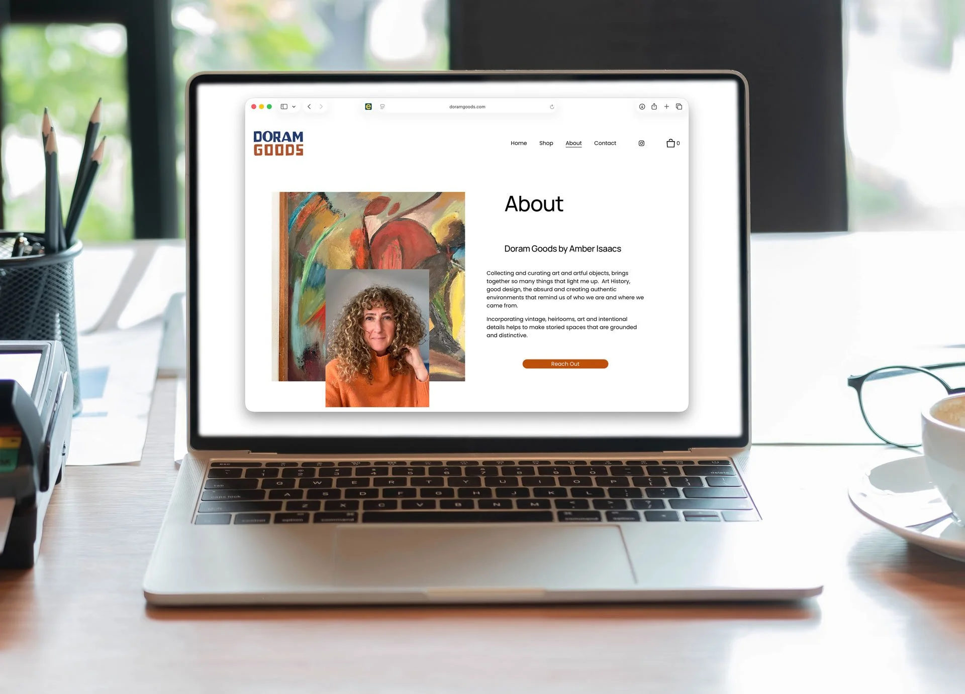Laptop screen displaying the 'About' page of Doram Goods website with a picture of a woman with curly hair standing in front of an abstract painting. Desk with office supplies and a cup of coffee nearby.