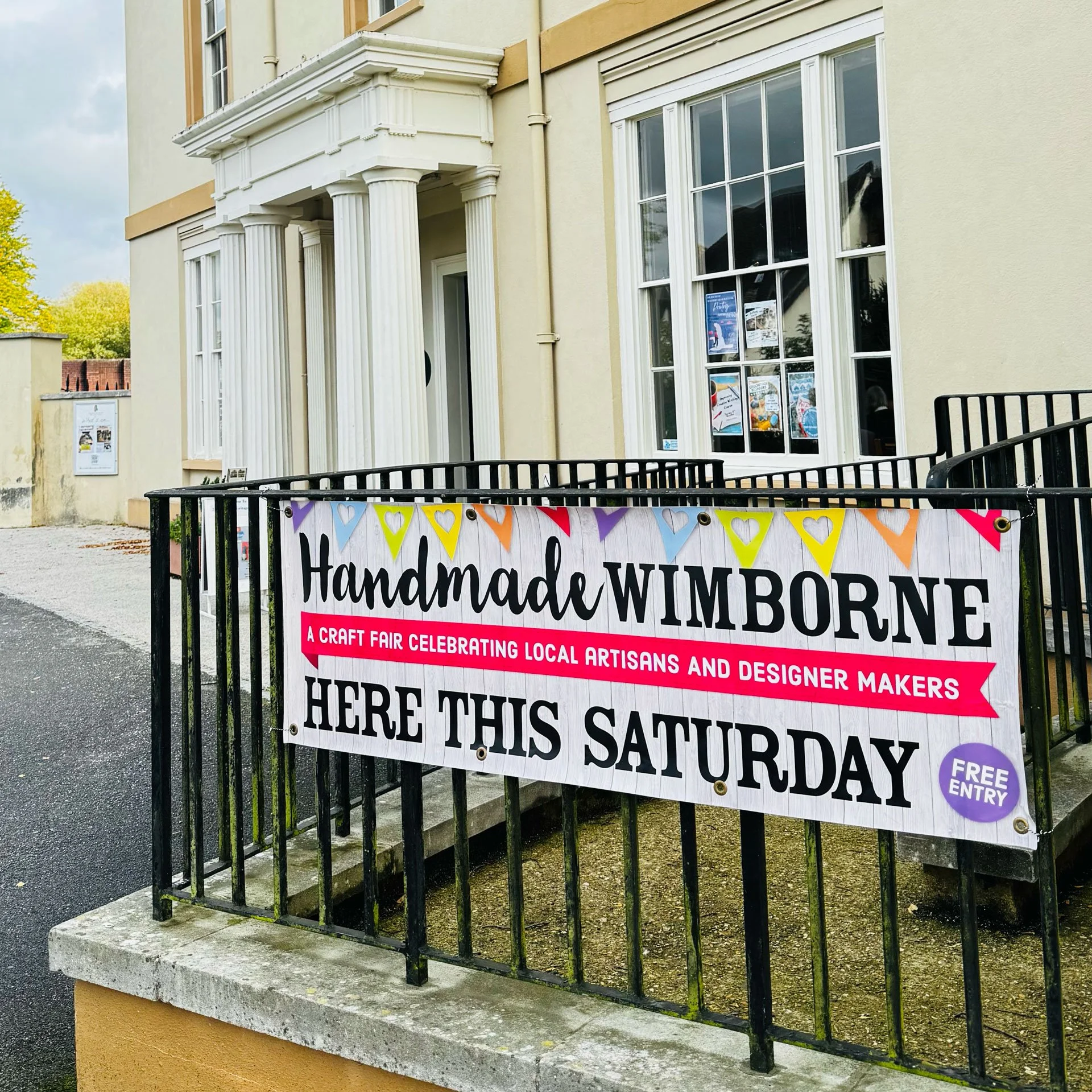 Banner advertising a craft fair highlighting handmade, Wimbome, local artisans and designers, with colorful heart-shaped decorations, attached to black metal fence outside a building with large windows and classical architectural details.