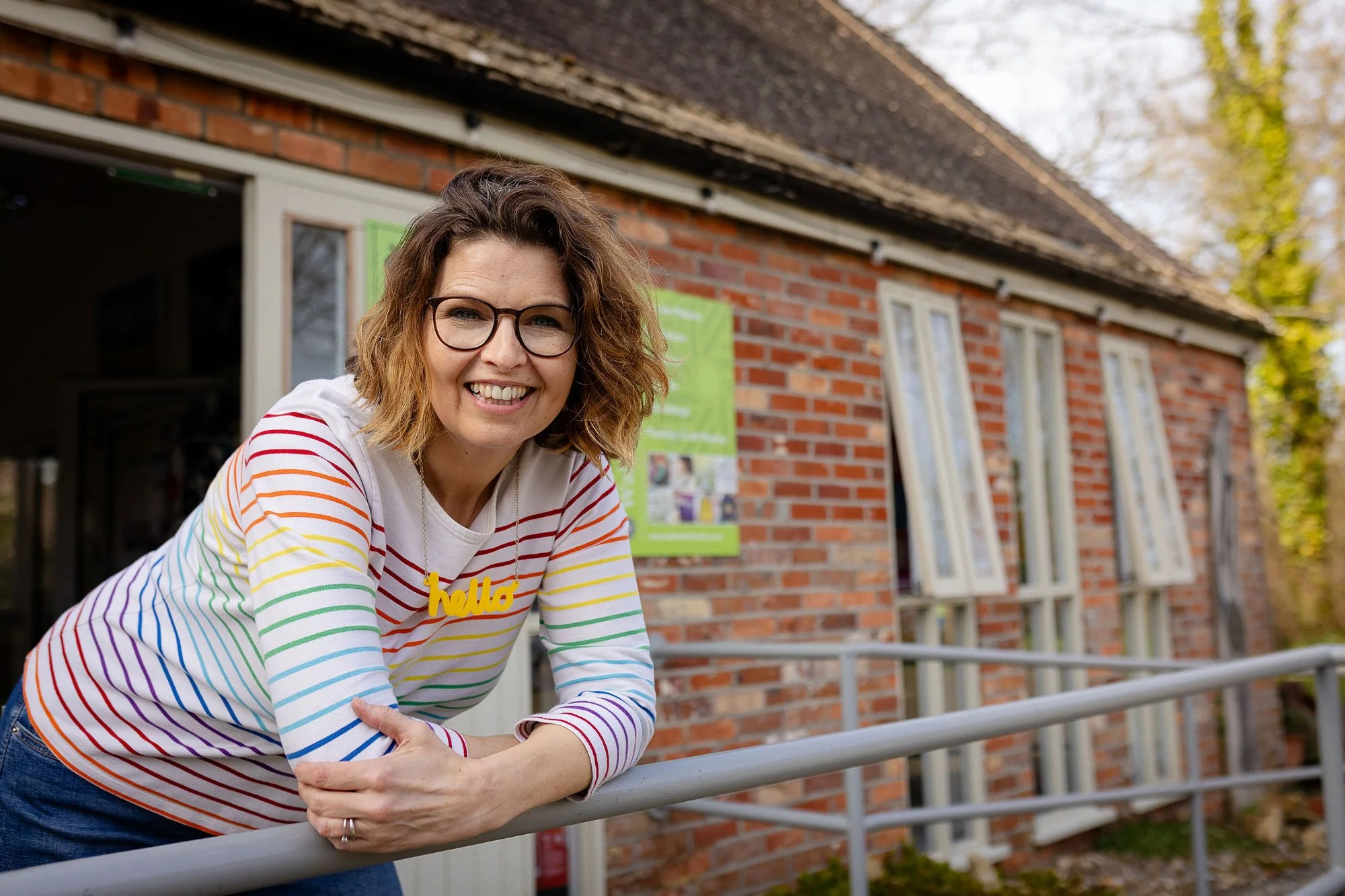 A woman with shoulder-length wavy hair, glasses, and a rainbow-striped shirt is smiling and leaning on a metal railing outside a brick building with green and white signs.