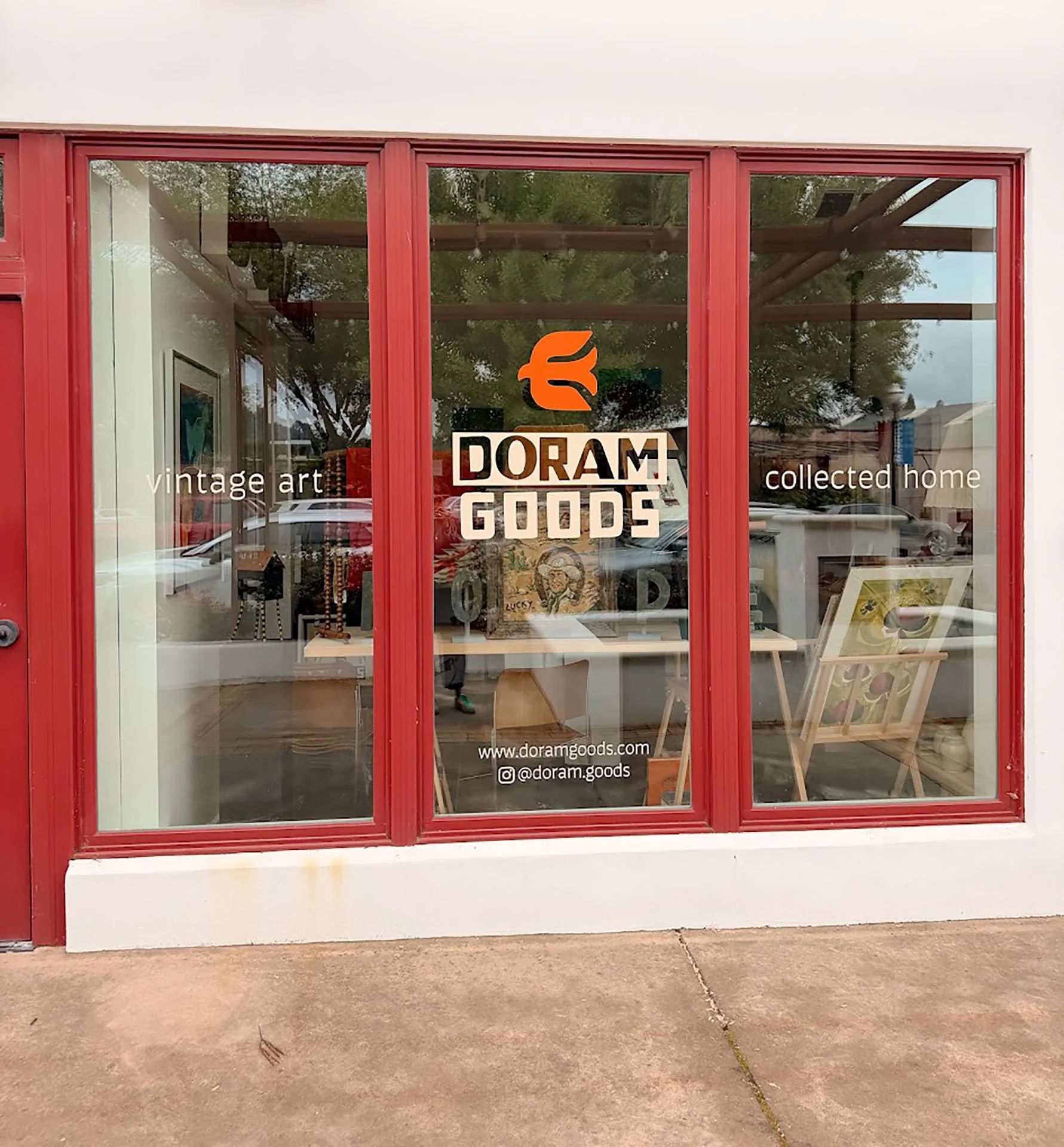 Front of a store with large glass windows and red framing. The store features signage with the words "vintage art" and "collected home," and the store's name "Doram Goods" with a logo above. Inside, some artwork and decorative items are visible.