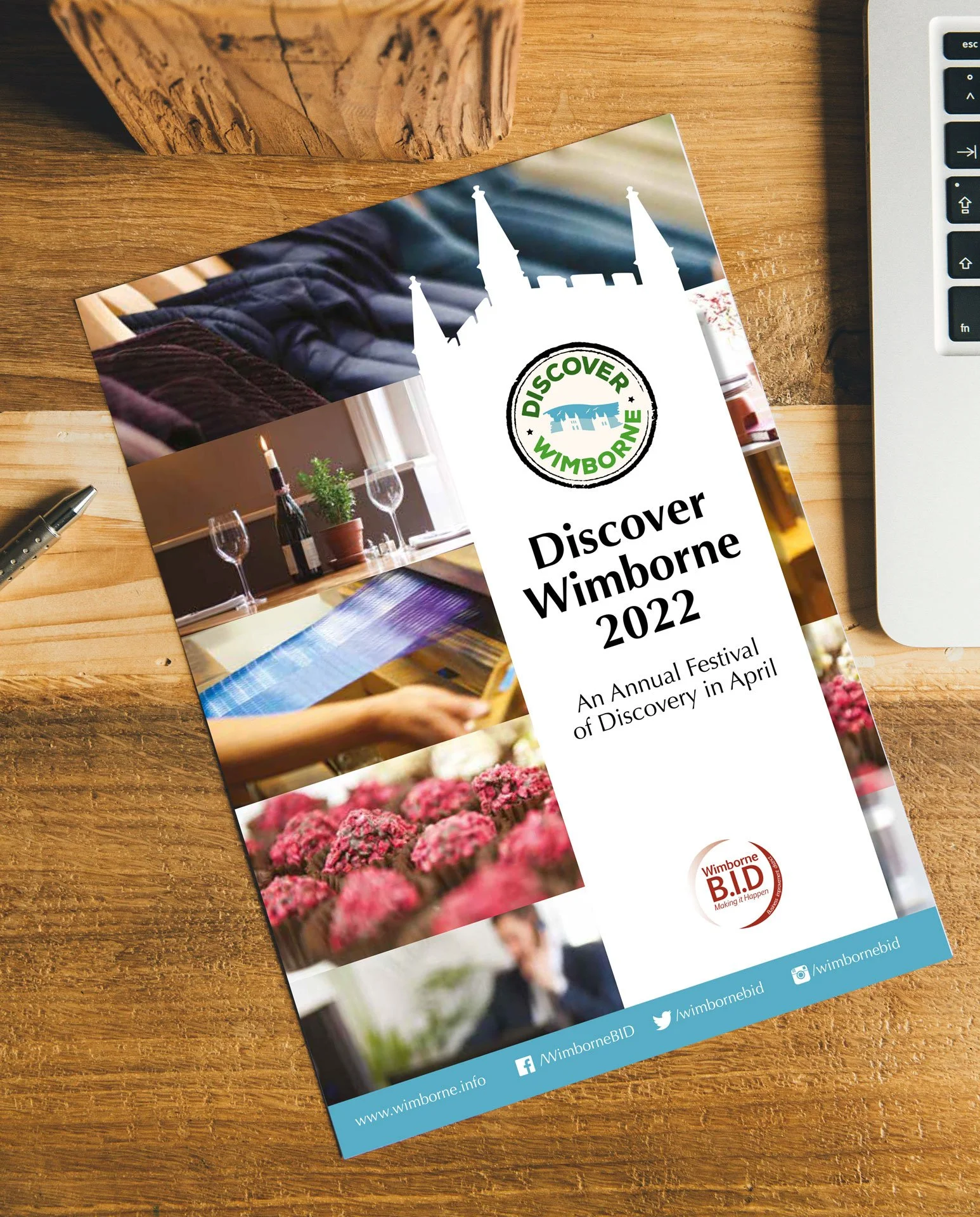 A brochure on a wooden table promoting the 2022 Discover Wimborne Festival featuring images of flowers, a wine glass, and a church silhouette, with social media icons and website information at the bottom.
