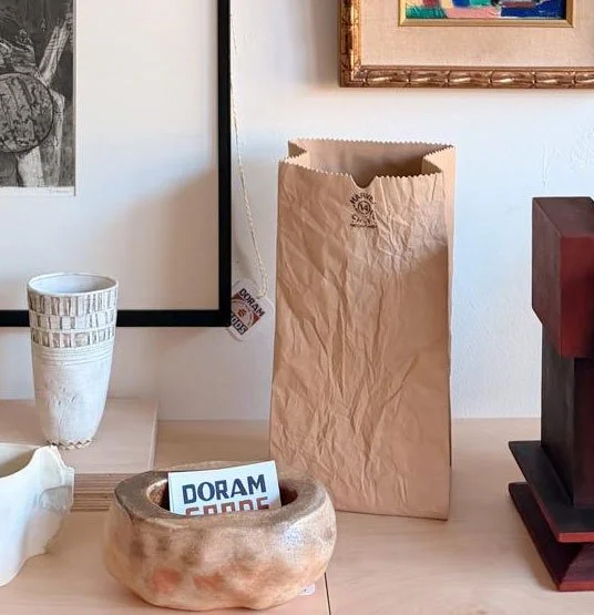Empty brown paper bag on a wooden surface with a white vase, ceramic bowl with a card inside, and a framed artwork on the wall behind.
