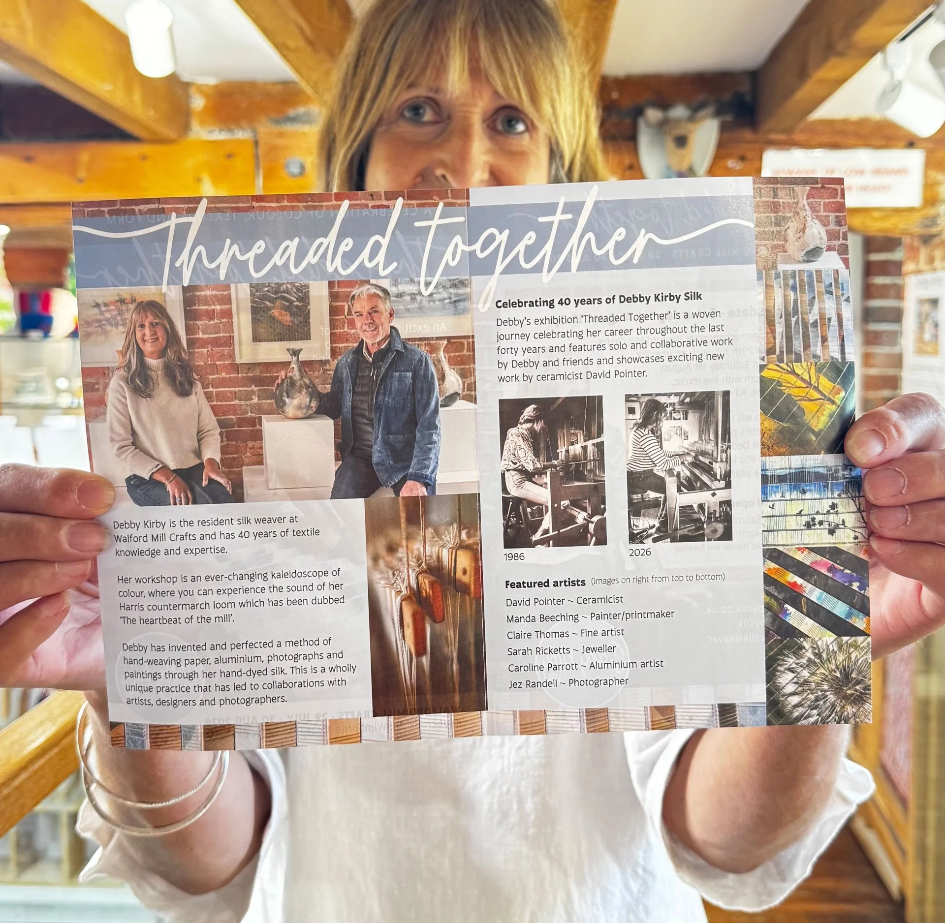 Person holding an exhibition flyer with images of two artists, Debby Kirby and David Pointer, and descriptions of their work, in a room with wooden beams and gallery artwork.