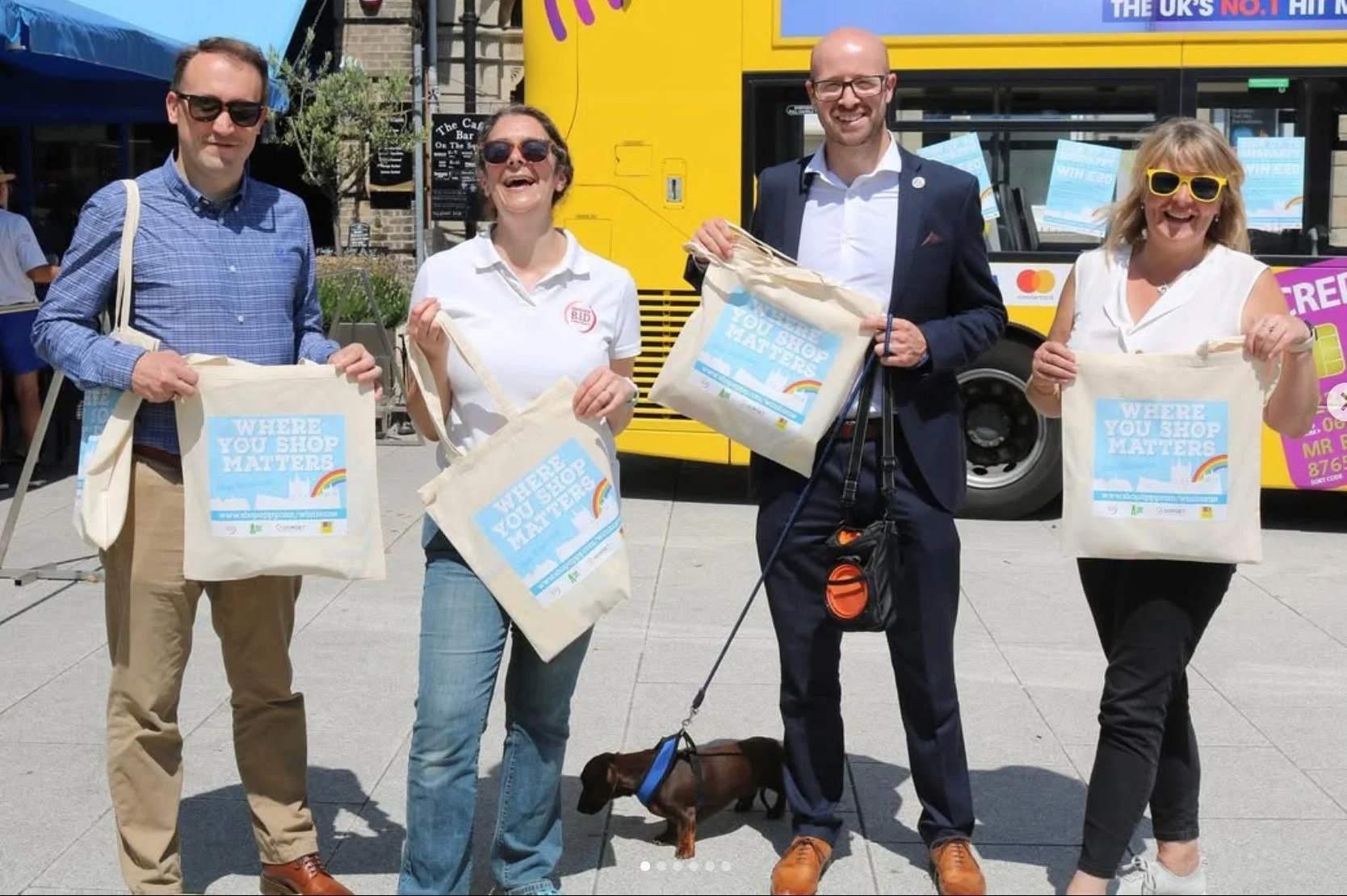 Four people standing outdoors holding tote bags that say 'Where You Shop Matters' in front of a yellow bus. They are smiling and wearing casual or semi-formal attire, and there is a brown dog on a leash in front of the person on the right.