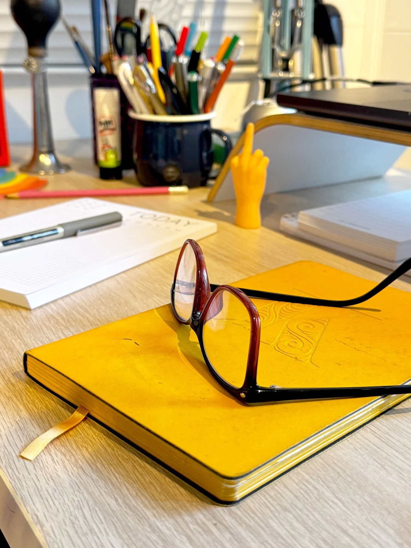 A yellow notebook with sunglasses resting on top, placed on a light wood desk. In the background, there are various office supplies including pens, pencils, and notebooks.