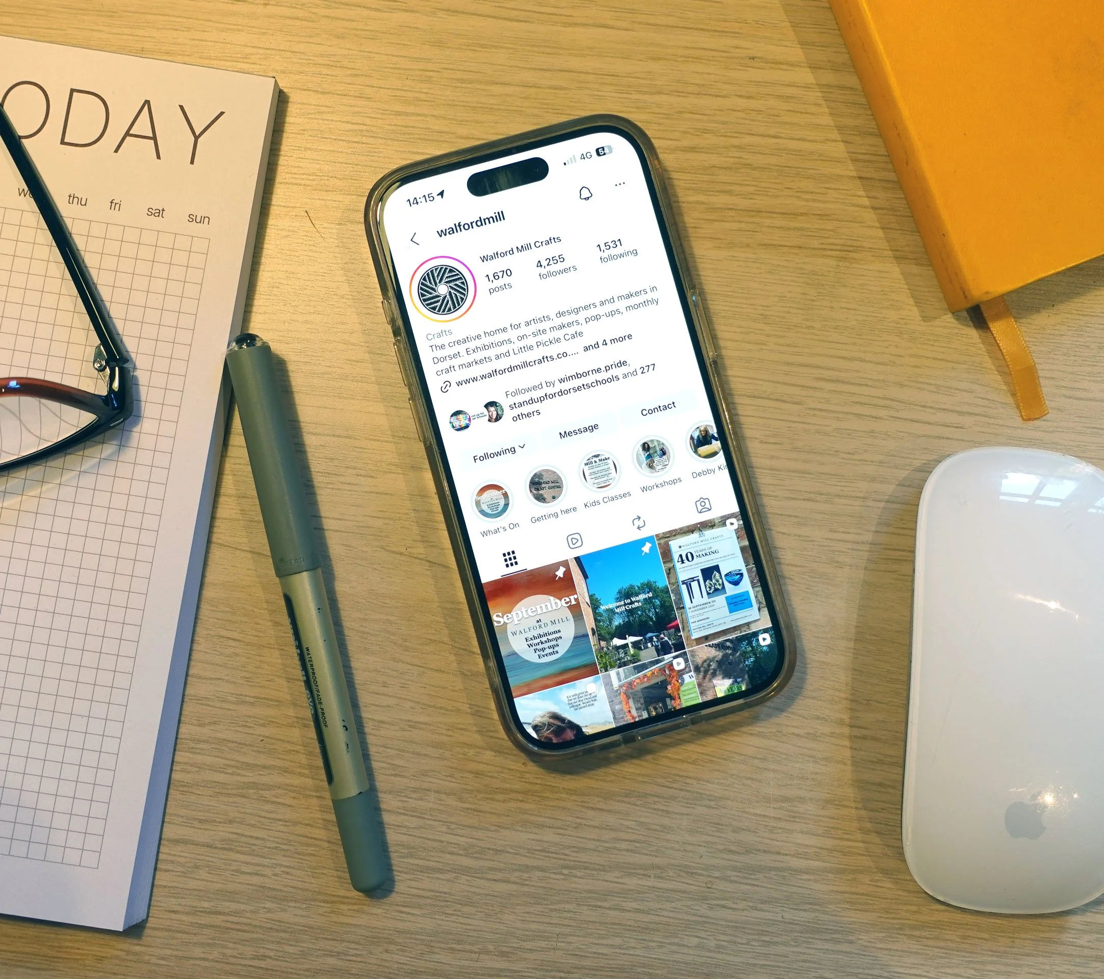 A smartphone displaying an Instagram profile for Walford Mill Crafts on a wooden desk, accompanied by a gray marker, a pair of glasses, a yellow notebook, a grid paper notepad, and a white circular object.
