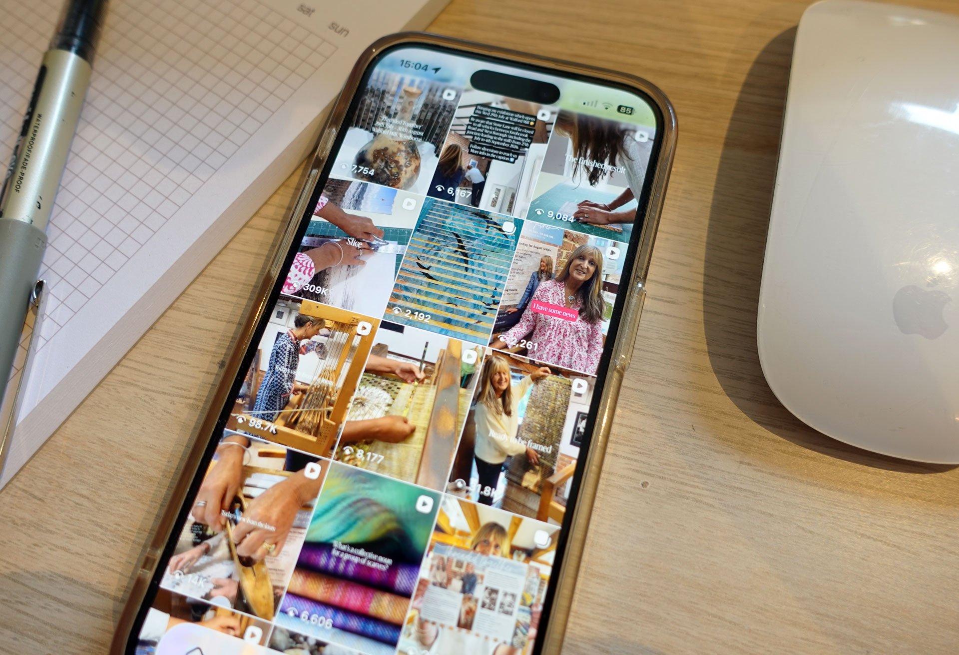 Smartphone on a light wooden desk displaying reels with a woman in a pink and white blouse, a woman knitting, and a child at a computer. Nearby are a white keyboard, a black and white marker, a notepad with a grid pattern, and a white computer mouse.