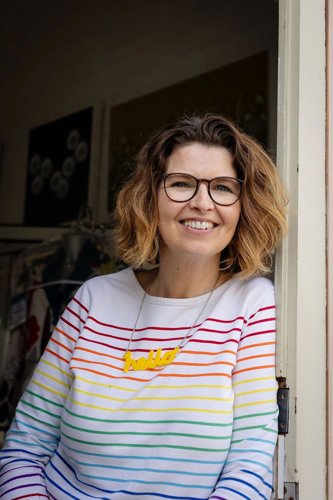 Smiling woman with wavy hair and glasses wearing a rainbow-striped shirt and a yellow 'hello' necklace, standing by a doorframe.