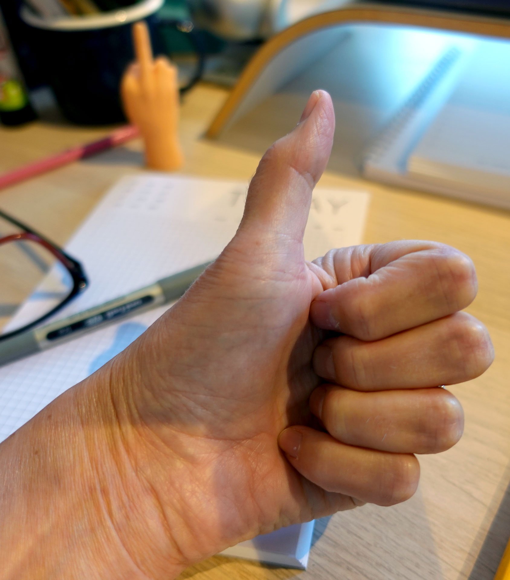 Close-up of a person's hand giving the OK gesture with a thumb and index finger touching, on a wooden table with various objects in the background.