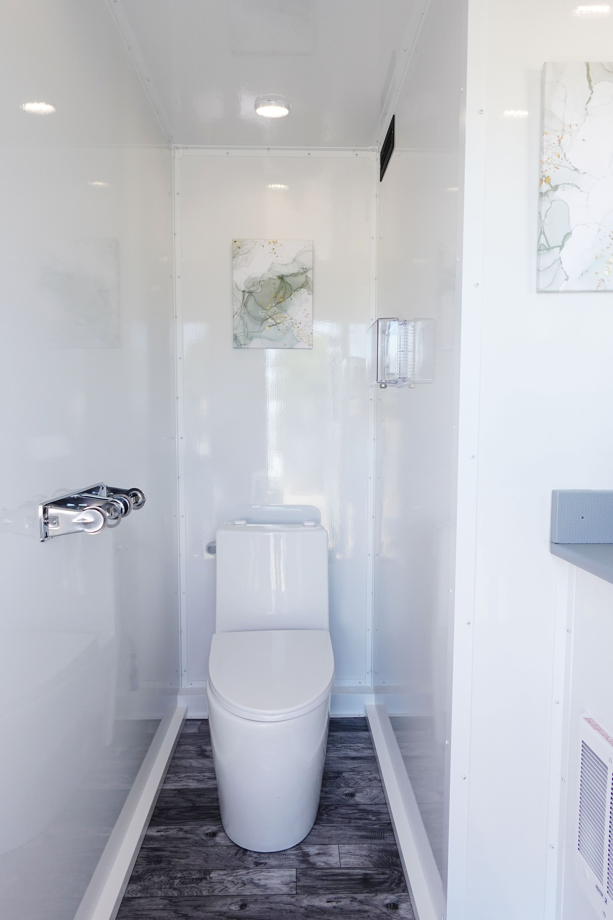 Small, modern bathroom with white walls and ceiling, featuring a white toilet, a small wall-mounted artwork with abstract design, a clear plastic tissue holder, and wood-patterned flooring.