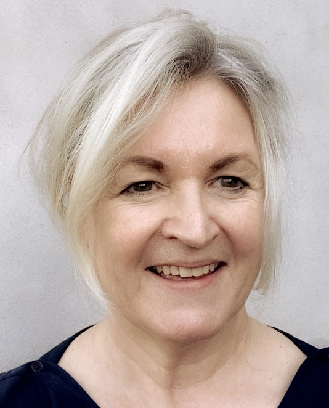Close-up photo of a smiling woman with short blonde hair, wearing a dark top, against a plain light-colored background.