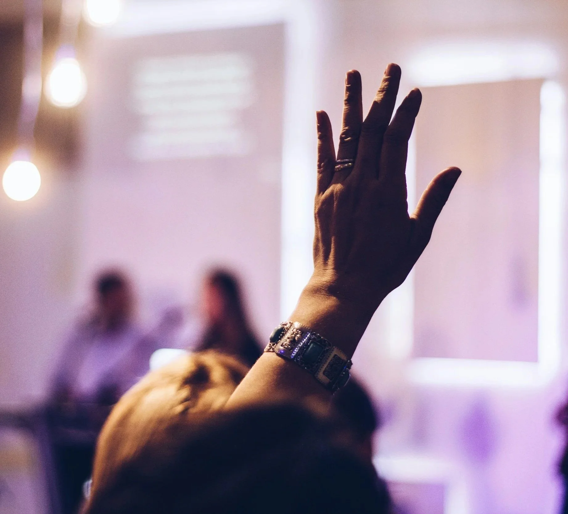 Close-up of a raised hand with a ring on the middle finger, wearing a watch, with a blurred background of a room with purple and pink lighting.