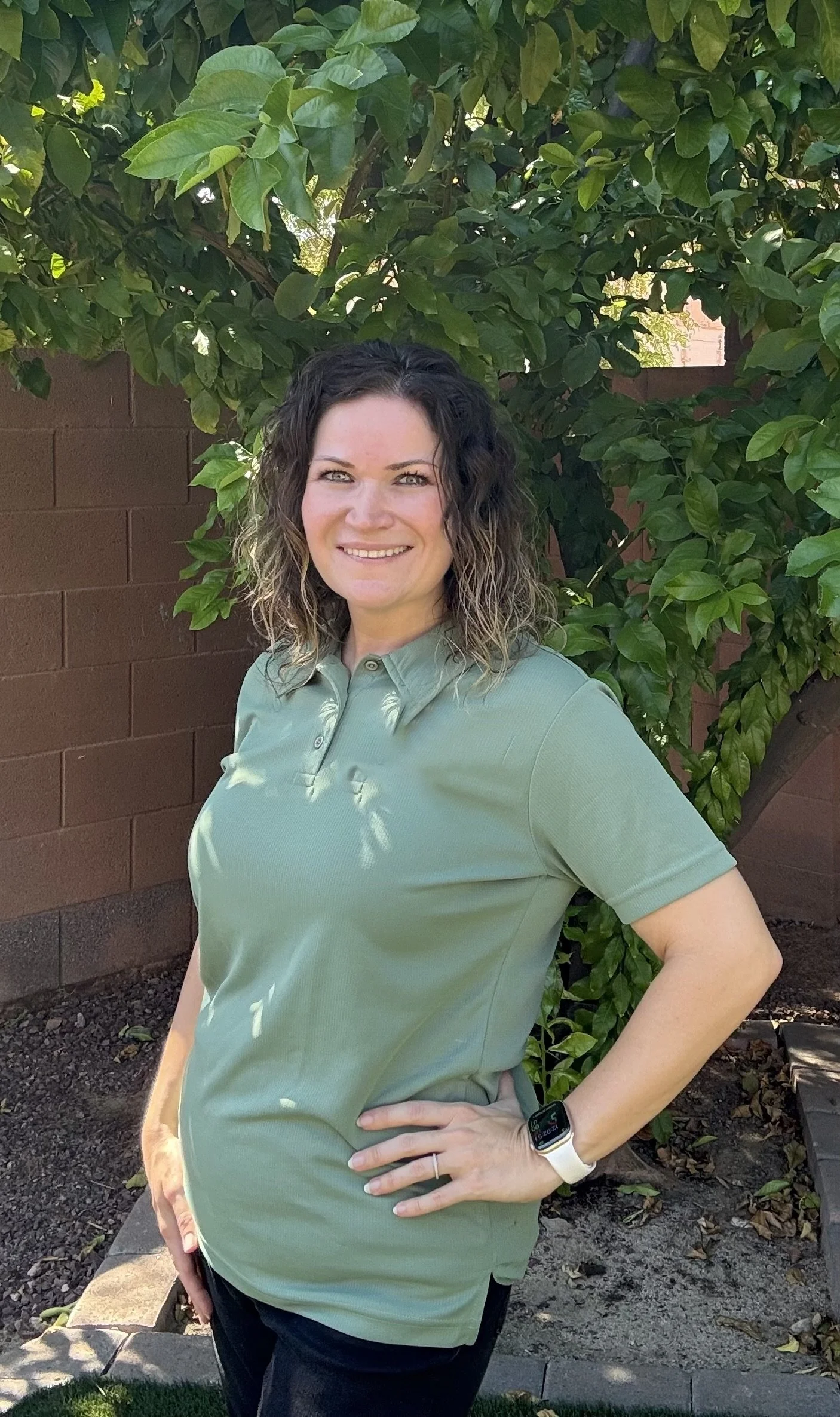 A woman with curly hair, smiling, wearing a green polo shirt, standing outdoors in front of green foliage and a brick wall.